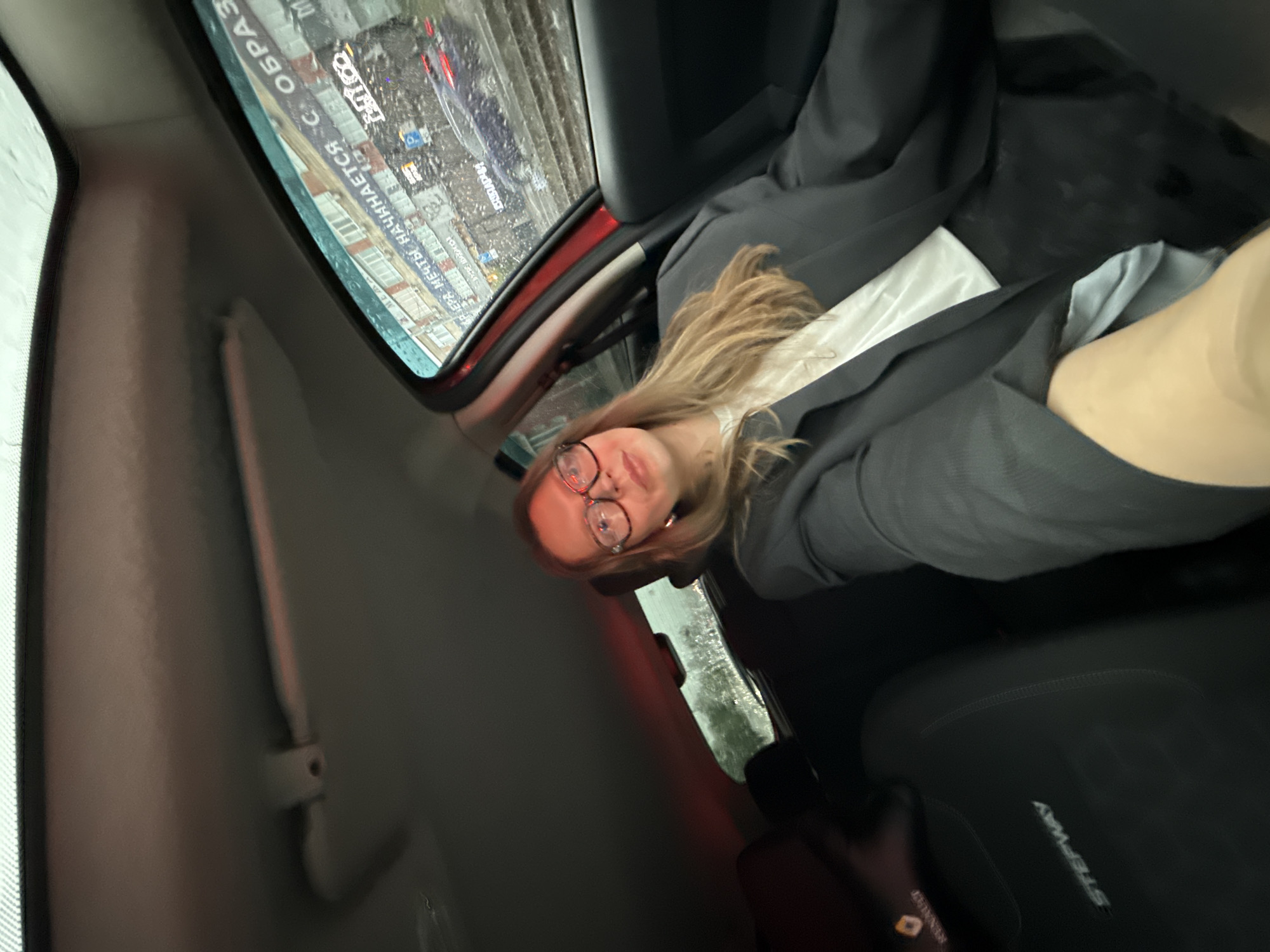 A young woman with blonde hair and glasses sits in the passenger seat of a car, wearing a black blazer over a white shirt. The car is presumably stopped, as there is a view of Izhevsk, Russia, out the rain-splattered window. Buildings and a street sign with the word "ОБРАЗ" visible in reflection are also visible in the view. The car seat is black and has "STEPWAY" stitched into the material.