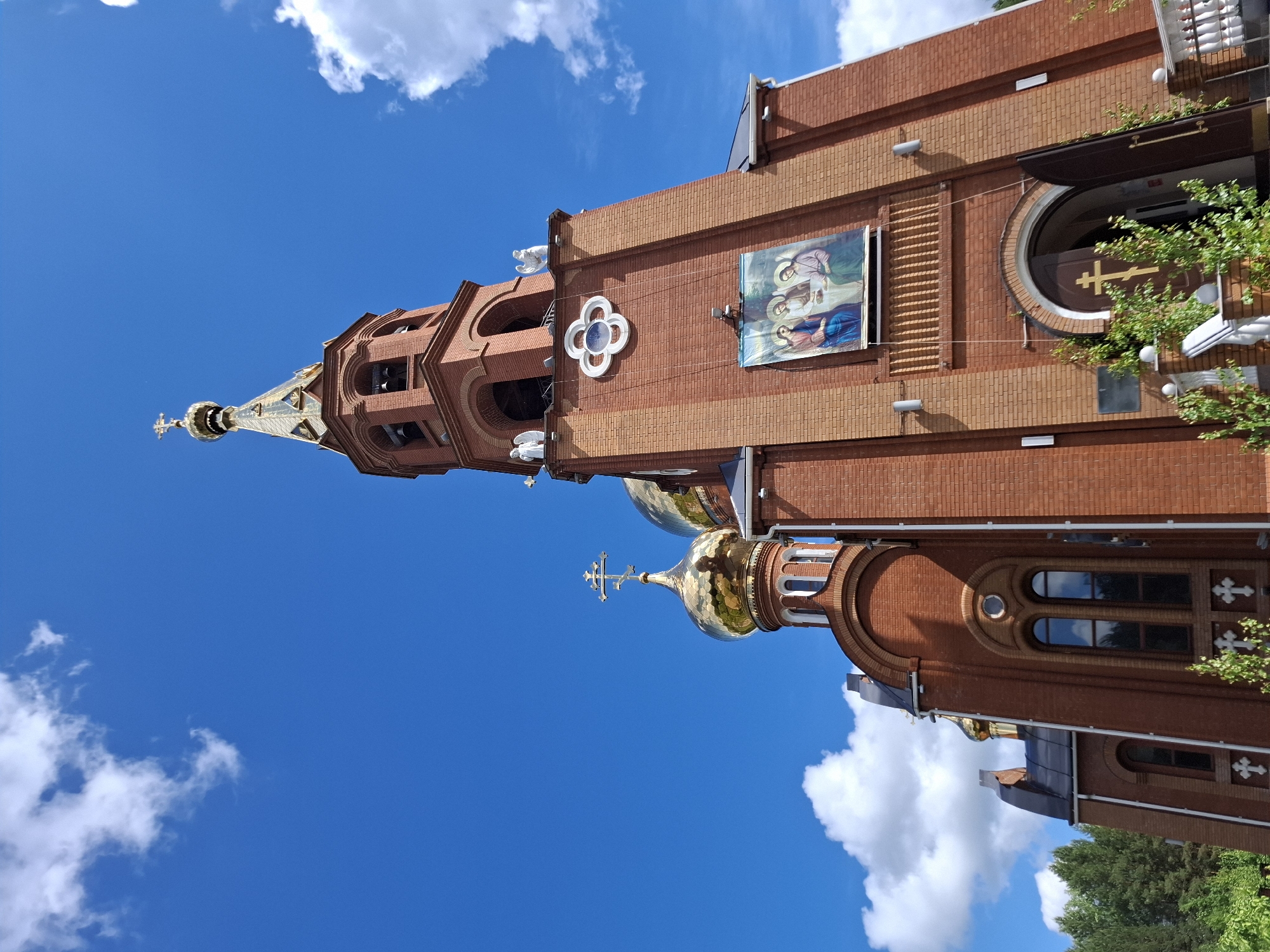 The image shows the exterior of a red-brick church building against a backdrop of a bright blue sky with white clouds. The church features a tall, ornate tower with a gilded dome and a cross at its peak. The walls of the church are adorned with religious iconography, including a framed painting of saints. The building has arched windows, decorative crosses, and a round entrance with a golden cross. The environment is urban, with glimpses of greenery. This is the church of the New Martyrs and Confessors of Russia, located in the city of Chaikovsky, Russia.
