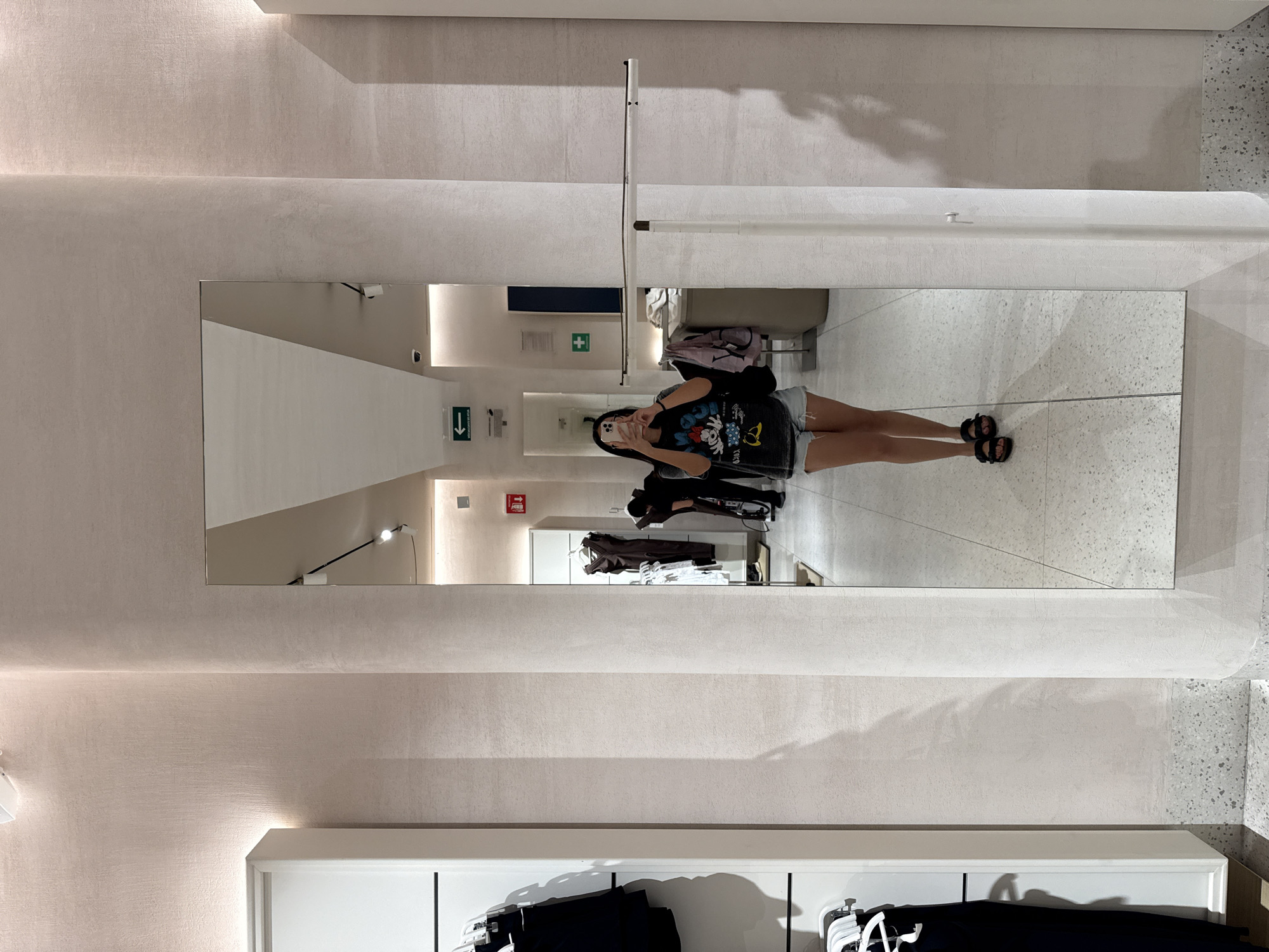 A person is seen in a full-length mirror within a retail environment. The location is identified as Puebla, Mexico. The individual is dressed in a graphic tee and shorts, holding a smartphone as if taking a photo of their reflection. The mirror's reflection reveals part of the clothing store interior, including racks of clothes, directional signage, and the store's overall aesthetic. The setting appears to be a clothing store's dressing room area.