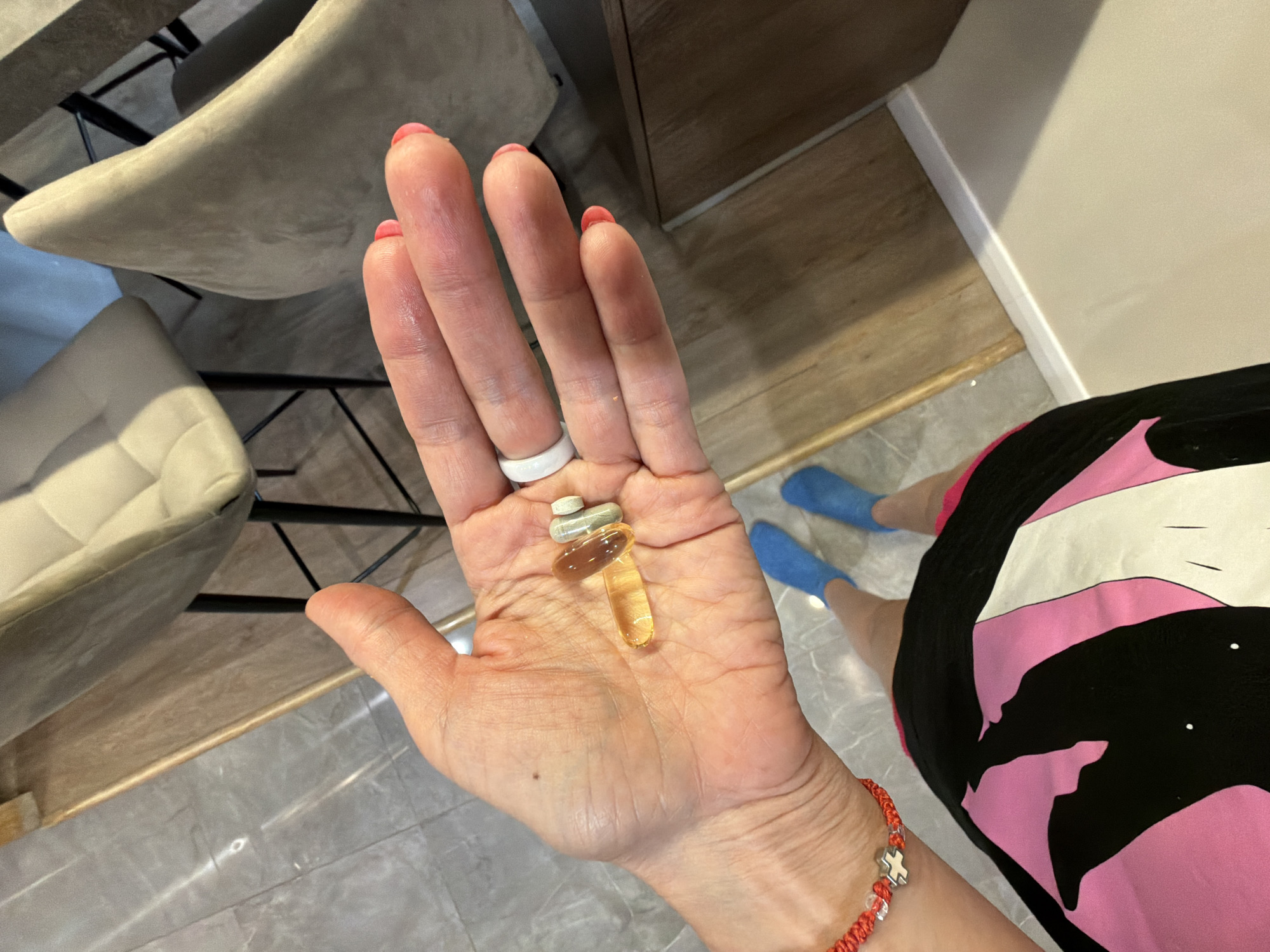 This overhead shot, set indoors in what is likely a private residence in Birobidzhan, Russia, captures a person's hand holding a daily regimen of supplements or medication. The hand, which appears to be female due to the pink nail polish and slender fingers, is adorned with a white silicone ring on the left ring finger and a red woven bracelet with a silver cross-like charm and clear beads on the left wrist.
In the palm of the hand are three pills: two light-colored, oblong tablets (one possibly white, the other light green or gray) and one translucent, amber-yellow gel capsule, likely a fish oil or vitamin supplement. The person's lower body is visible in the background; they are wearing a black garment with bold, abstract pink and white patterns, and light blue slipper-socks on their feet.
The setting appears to be a well-lit room during the daytime. The floor beneath the person consists of large, light gray tiles, transitioning to a strip of light wood flooring along a wall with a white baseboard. In the background, two modern chairs with light-colored, possibly upholstered seats and dark metal frames are visible, along with a partial view of a wooden cabinet or furniture piece. No text is visible in the image. The scene suggests a routine moment of health maintenance.