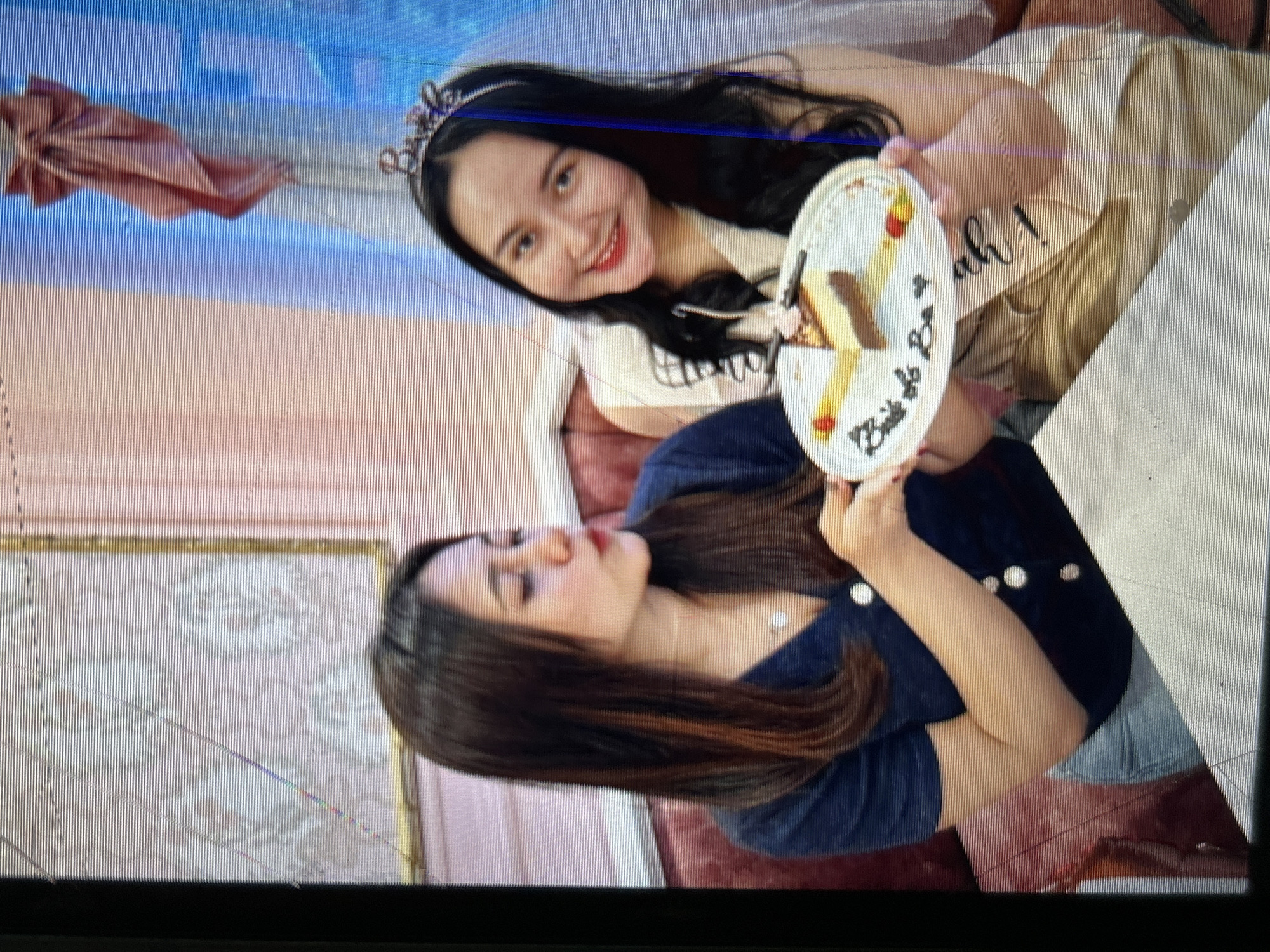 Two young women are pictured in a bright, indoor setting, possibly a cafe or event space in Depok, Indonesia. The woman on the left, wearing a dark blue V-neck top and light blue jeans, is holding a white plate with a slice of cheesecake on it. The cheesecake has decorative elements and writing on the plate that appears to say "Bride to Be." She is looking to her right with her lips pursed. The woman on the right, who is wearing a light-colored sleeveless top and a sash that reads "Bride," is smiling widely and looking at the camera. She is wearing a tiara on her head. The background features a pink wall with a diamond pattern and a sheer pink curtain. A large pink bow is visible in the upper right corner of the image.