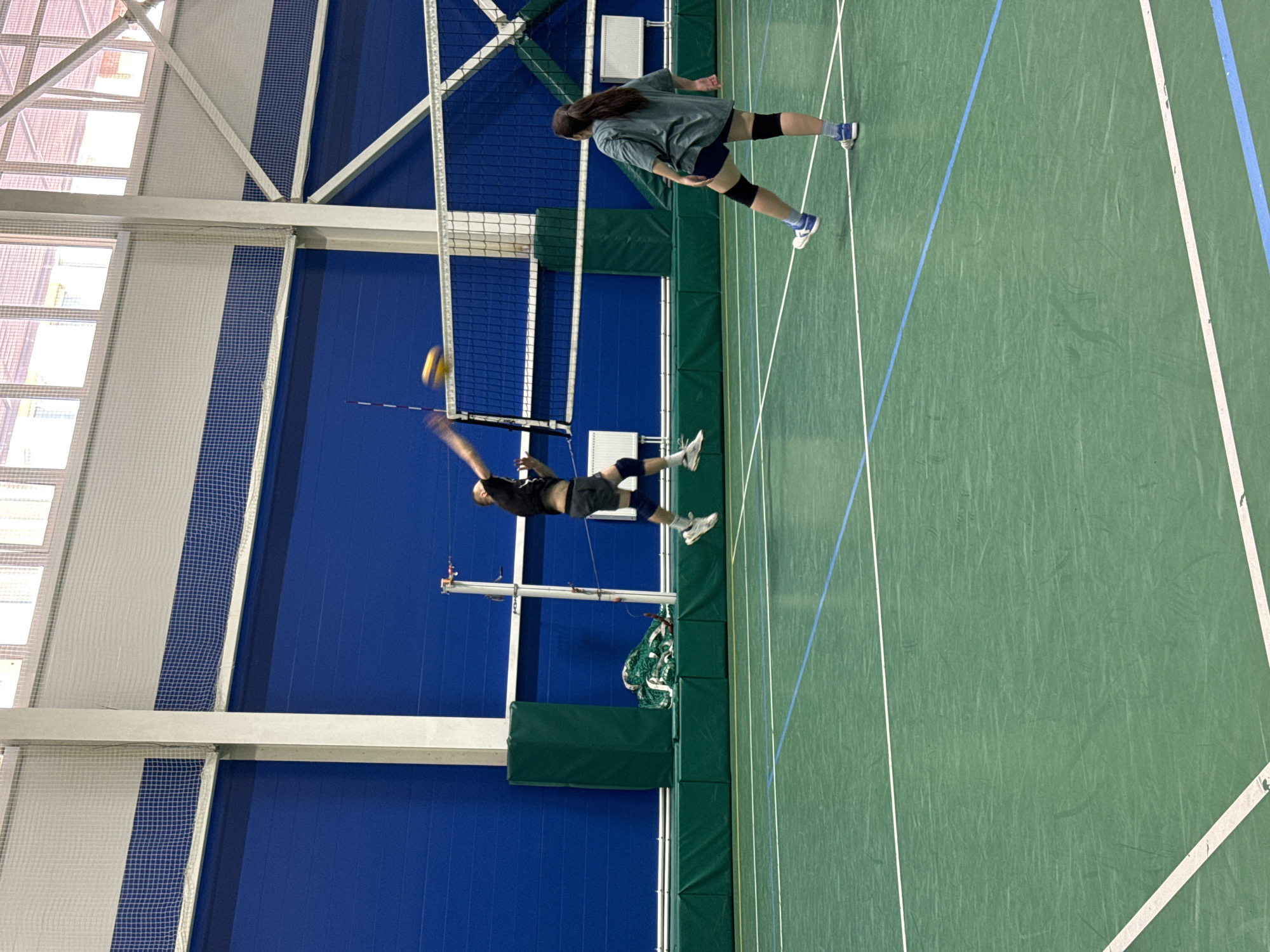 This image depicts an indoor volleyball match taking place in a gymnasium. Two individuals are actively engaged in play, with one player in mid-air, appearing to spike the ball over the net, while another player is positioned on the opposite side of the court, facing away from the camera. The court is marked with white and blue lines, and a volleyball net divides the playing area. The walls of the gymnasium are a deep blue, and the ceiling features a grid-like structure with white netting. The floor is a green athletic surface. The presence of gym equipment and the overall structure of the building suggest it is likely a sports facility within Saint Petersburg, Russia.