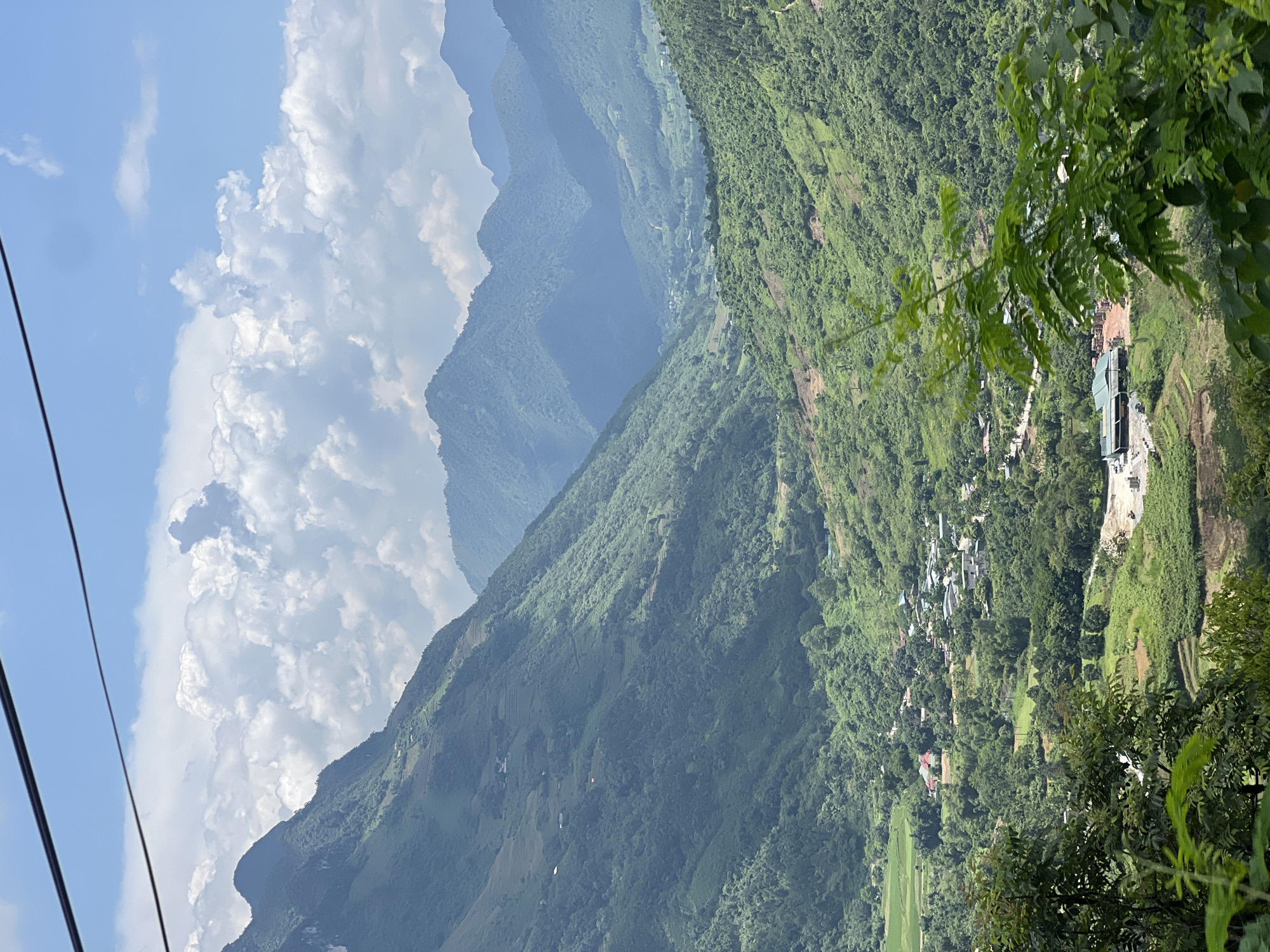 This is a vertical, eye-level, medium shot of a lush valley in Minh Tân Commune, Vietnam, under a bright, partly cloudy sky. The foreground features lush green trees and foliage obscuring the lower portion of the frame. The middle ground reveals a vibrant green valley dotted with houses, some with red roofs, and agricultural fields. Terraced fields climb the hillsides, showcasing human cultivation amidst the natural landscape. In the background, successive layers of majestic, mist-shrouded mountains recede into the distance, their slopes covered in dense vegetation. The overall impression is one of a serene and picturesque rural landscape.