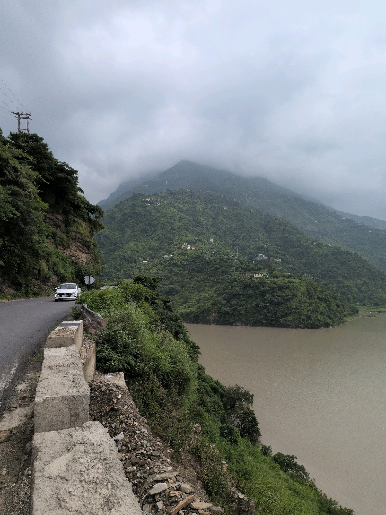 A white sedan drives on a paved road that curves along a steep, forested mountainside. The road is bordered by a concrete barrier and a steep drop to a large, murky body of water. The mountains in the background are covered in lush green vegetation and dotted with scattered houses. The sky is overcast with clouds, and the overall mood of the image is serene and natural. The scene depicts a road trip through a mountainous region, likely in a rural area. The presence of the power lines on the left suggests that the area is inhabited and connected to infrastructure.