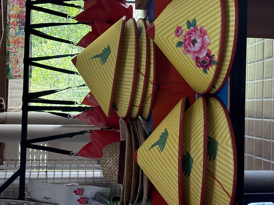 This image captures a market stall display in Takua Thung, Thailand. The foreground is dominated by traditional conical hats and other headwear arranged on a red and blue display surface. On the left, a stack of at least four woven straw hats with wide brims and dark red bands is visible, topped by a hat with a tightly woven basket-weave pattern. Adjacent to these are three yellow, woven conical hats with red trim, each adorned with a green fern-like leaf motif, stacked vertically. To the right, a central stack consists of four identical yellow, woven conical hats with red trim and green fern-leaf motifs, some revealing red chin straps with black toggles. Further to the right, a single yellow, woven conical hat with red trim is prominently displayed, featuring a large pink floral design. Above the hats, a black horizontal bar spans the frame, from which multiple bright red, waterproof dry bags with black shoulder straps are suspended. The background reveals an outdoor setting with lush green foliage visible through an open space, suggesting a natural environment beyond the display. Architectural elements like pillars and beams are present above the foliage, and a colorful banner with partial Thai script, including "ตุลาคม" (October), is visible in the upper background. The lower foreground shows a textured floor or counter surface composed of light brown, rectangular tiles. No individuals are depicted in the scene.