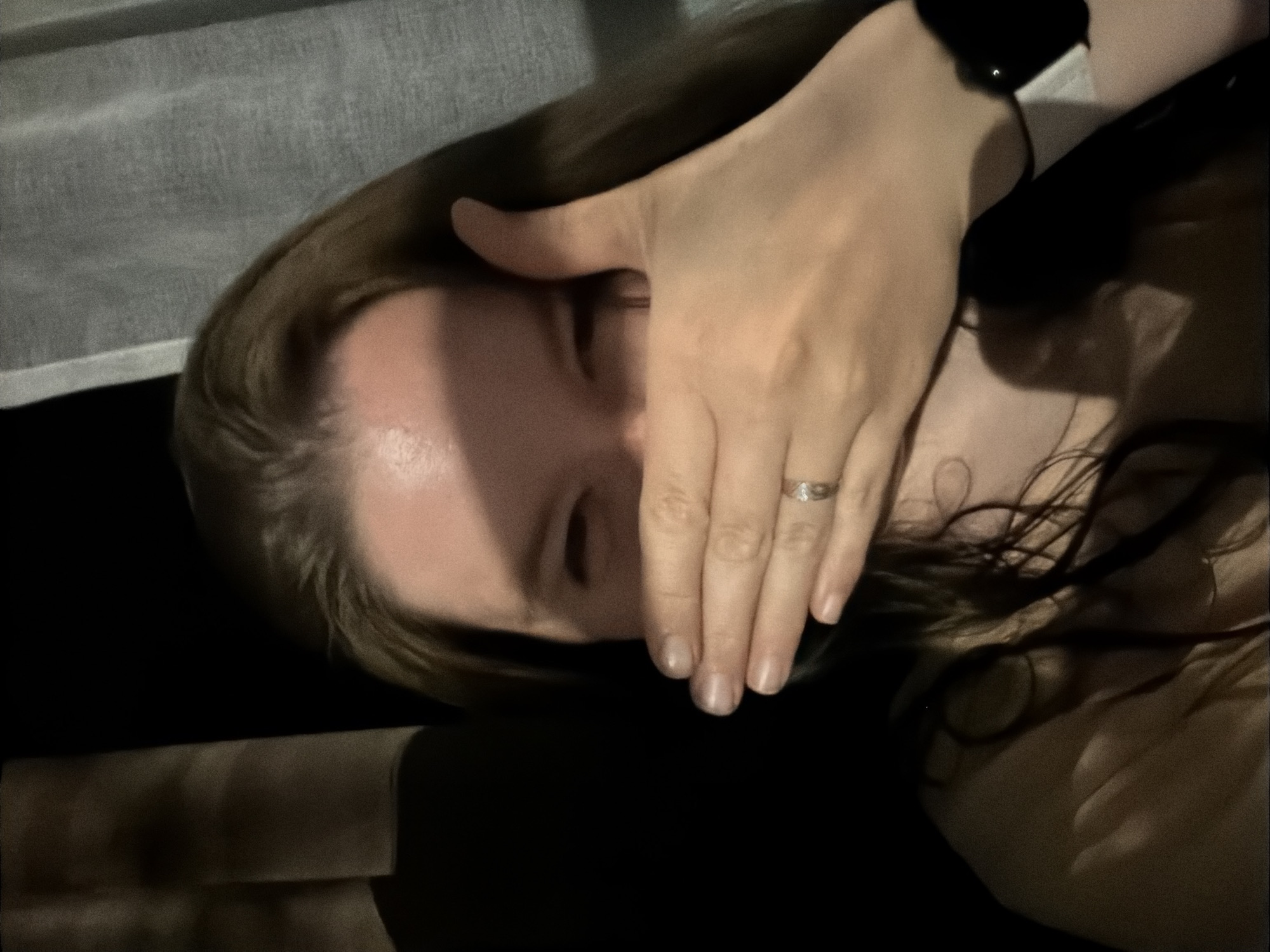 A person with long, light brown hair is lying down with their left hand covering their face. The person wears a ring on their finger. A black object, possibly a watch or fitness tracker, is visible on the person's left wrist. The background includes a partially visible white curtain. No location in the city of Staraya, Russia is discernible from the image.