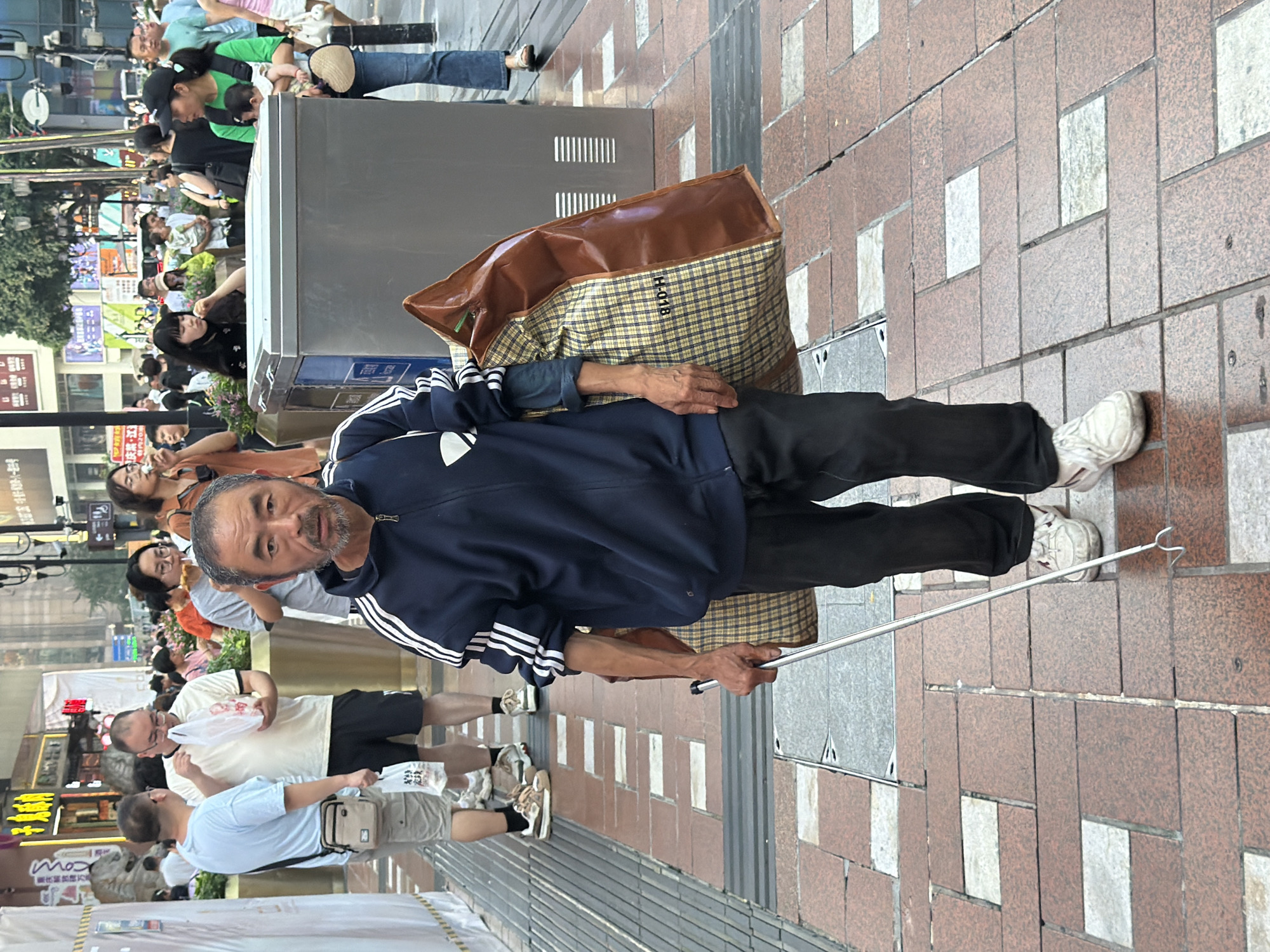 A middle-aged East Asian man, who appears to be visually impaired, is walking down a paved city street in Yuzhong District, China. He is holding a silver-colored walking cane with a curved tip in his right hand. He is wearing a dark blue tracksuit jacket with white stripes on the sleeves, dark pants, and white sneakers. The man is carrying a large, plaid duffel bag with brown leather trim over his left shoulder. He has a stern expression on his face and a gray beard. The street is lined with shops and other pedestrians. The lighting suggests it is daytime, likely overcast.