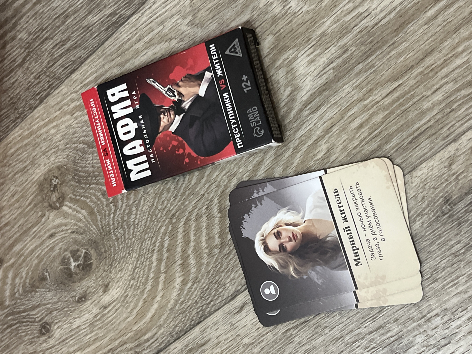 This image shows a top-down view of a game box and cards laid out on a wooden surface. The game box is red and black with the title "МАФИЯ" (Mafia) prominently displayed, along with the subtitle "НАСТОЛЬНАЯ ИГРА" (Board Game). The box depicts a man in a suit and fedora holding a pistol. Text on the box indicates "ПРЕСТУПНИКИ VS ЖИТЕЛИ" (Criminals vs. Citizens), "SIMA LAND", and "12+". To the left of the box, a stack of cards is visible. The top card features an illustration of a woman with blonde hair and the title "Мирный житель" (Peaceful Citizen). Below the title, text describes the character's task: "Задача – ночью закрыть глаза, а днём участвовать в голосованияи" (Task - close eyes at night, and participate in voting during the day). The wooden surface appears to be a floor with a wood grain pattern. The scene is indoors. There are no people in the image. The location is not specified as the city of Дачное, Russia, as this information is not present in the image.