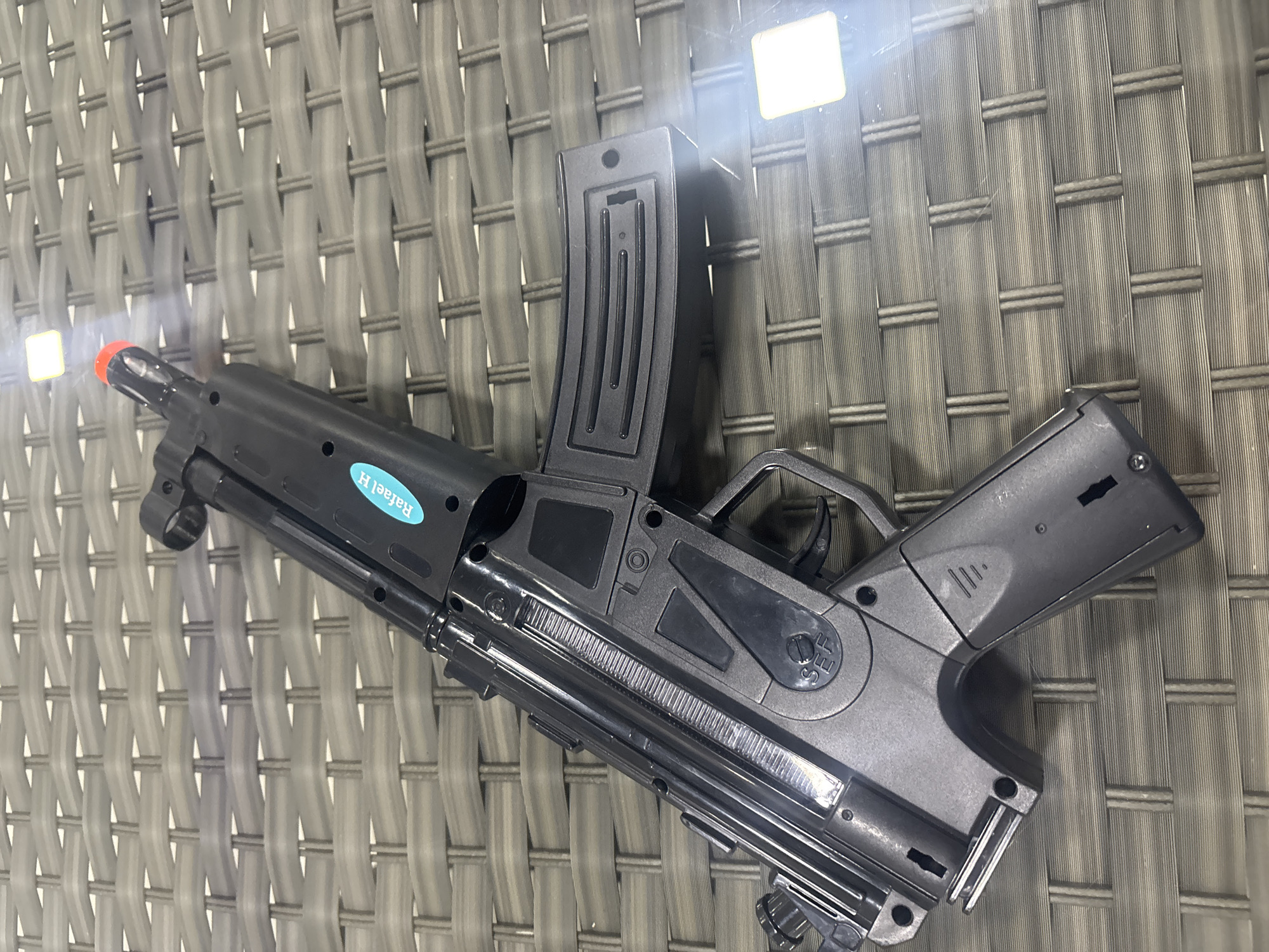A black toy assault rifle, resembling a Heckler & Koch MP5, rests diagonally on a gray wicker-patterned surface. The toy gun has a magazine inserted, a red-tipped barrel, and a blue sticker with white text that appears to read "Rafael H". The scene is illuminated by artificial light sources that create bright rectangular reflections on the surface. The background is a uniform gray wicker pattern. No individuals or specific city landmarks are visible. The image appears to be taken indoors or on a covered outdoor surface, as indicated by the reflections and lack of natural sunlight. The setting is not identifiable as Dubai, United Arab Emirates.