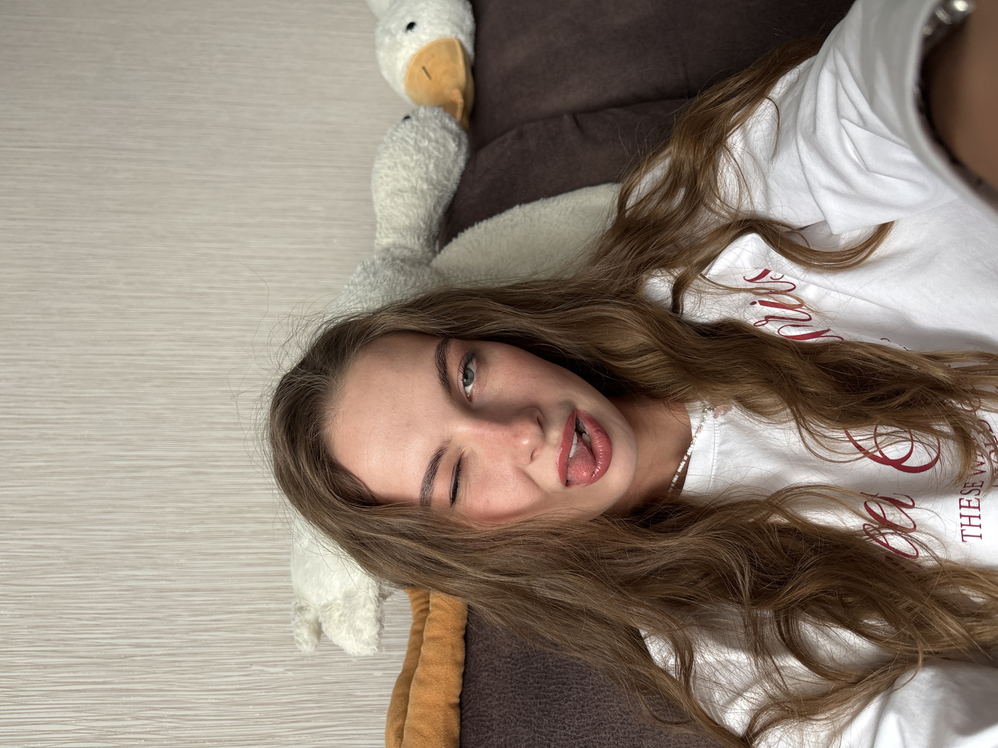 A young female individual is depicted in a self-portrait taken indoors in Perm, Russia. She has long, wavy brown hair that cascades over her shoulders and chest. She is wearing a white long-sleeved top with red cursive lettering, partially visible as "THESE W" and "PRIUS" (or similar) on the chest. A white and pink beaded necklace is visible around her neck. Her right eye is closed in a wink, and her left eye is open, revealing a light-colored iris. Her mouth is slightly open, with her tongue extended forward. Her lips are coated with a glossy, reddish-pink product.
She is positioned against a dark brown upholstered couch. Behind her, on the couch, are several plush items: a white stuffed duck with an orange beak is visible behind her right shoulder, and a larger white textured plush object is situated behind her left shoulder. An additional orange-brown soft item or blanket is on the couch to her left. The background consists of a light beige or off-white wall with a subtle vertical striped texture. A portion of her right arm and wrist is visible in the bottom right corner, consistent with holding a device to capture the image.