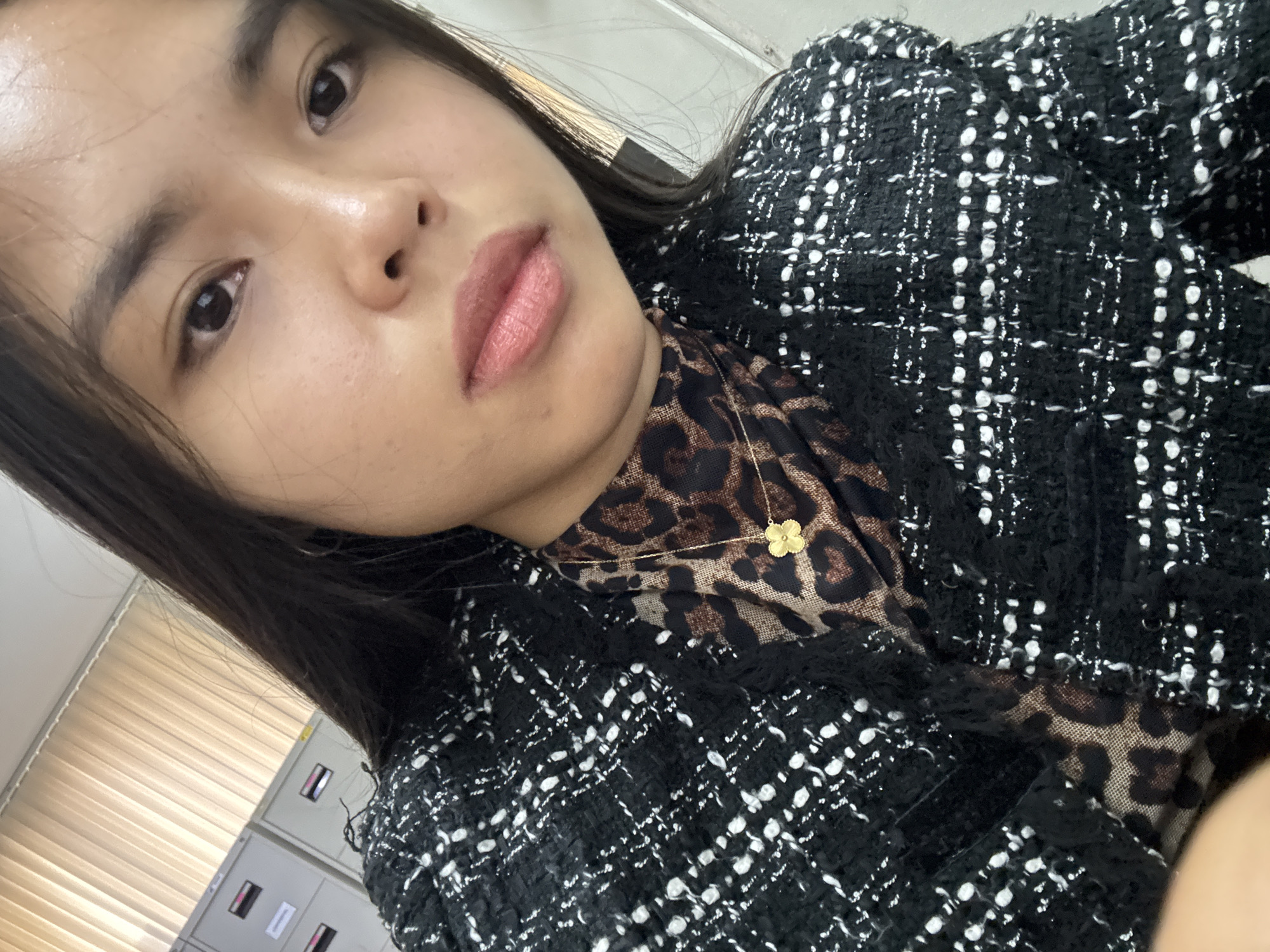 A young woman with long, dark hair and light brown eyes is looking directly at the camera. She is wearing a black and white tweed jacket over a leopard-print turtleneck. A gold necklace with a clover-shaped pendant rests on her chest. The background shows filing cabinets and vertical blinds, suggesting an indoor office environment. There are no overt indications of the specific location within Davao City, Philippines, beyond the general context of an office setting.