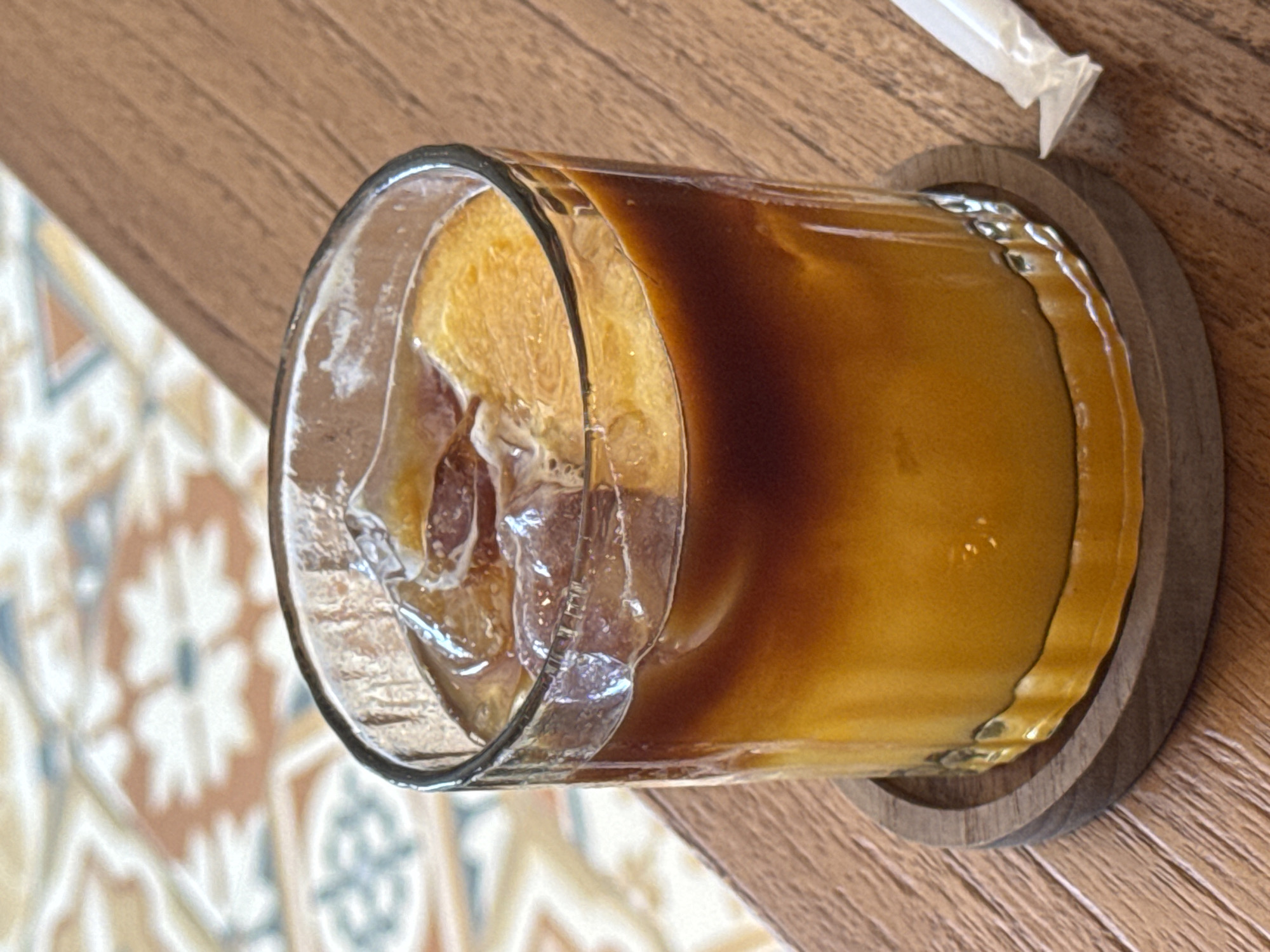 The image shows a glass of iced coffee with a straw, likely an iced latte or similar beverage. The glass sits on a wooden coaster on a table. Part of the table and decorative tilework are visible in the background. Based on the location information (Ungasan, Indonesia), it is likely the drink was purchased at a café or restaurant. No people are visible. The focus is the drink and the surrounding table setting.