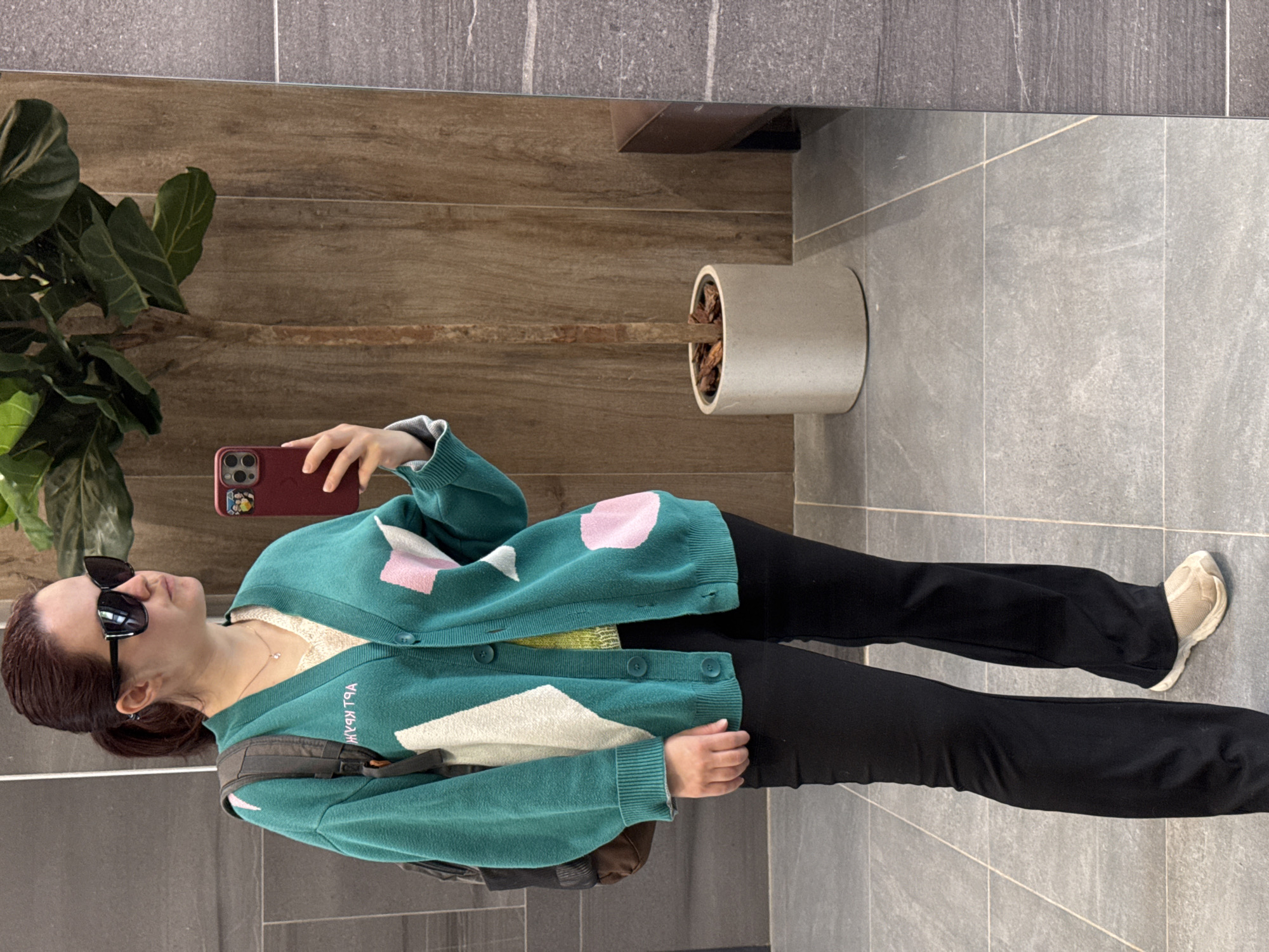 A woman stands in what appears to be an indoor space, possibly a building lobby or a hallway. She is taking a selfie in a mirror using a red smartphone. She is wearing sunglasses, a teal cardigan with white and pink geometric shapes, a light-colored top underneath, black pants, and off-white sneakers. She also has a backpack on. A potted plant is visible to the left of the woman, and another planter is seen near the mirror's edge. The background includes brown wooden wall panels and gray tiled flooring and walls. The location is not identified in the image.