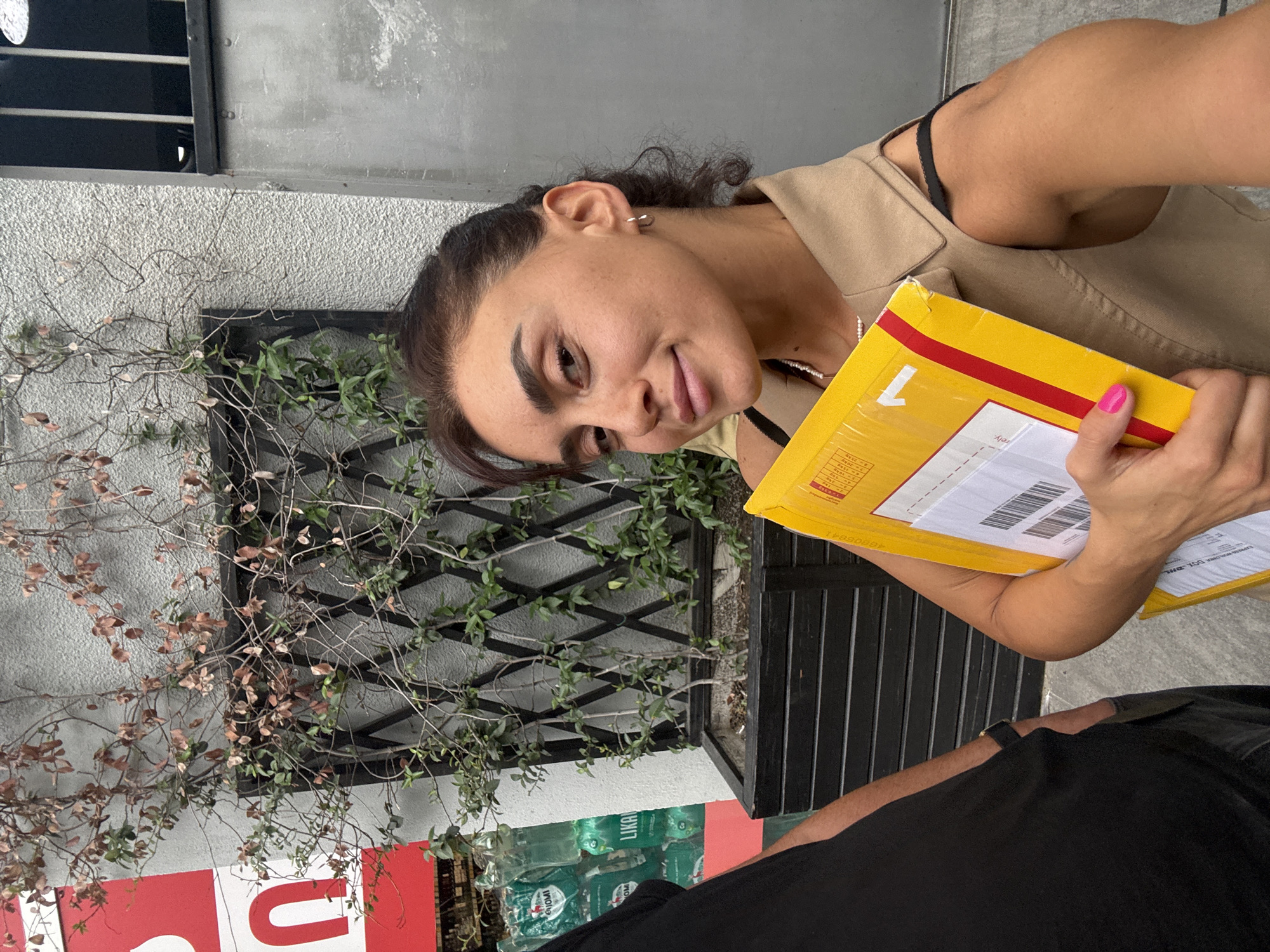 A young woman is standing outside on a sidewalk, holding a yellow package. She is wearing a tan vest over a black top. Her hair is pulled back, and she is wearing small hoop earrings. A man in a black shirt is partially visible on the left side of the frame. In the background, there is a building with a textured wall, a metal grate with ivy growing on it, and a wooden planter box. The woman's fingernails are painted bright pink. The woman is looking towards the camera and smiling. The scene appears to be in an urban environment.