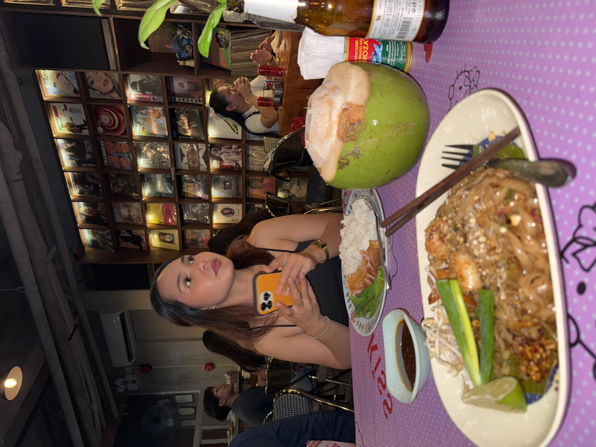 The image captures a lively indoor scene at a restaurant, possibly in Davao City, Philippines, as suggested by the context. In the foreground, a woman is holding an orange smartphone, her attention seemingly directed elsewhere. She is seated at a table covered with a purple tablecloth adorned with a pink "Kiss" pattern and small white polka dots. On the table, a plate of Pad Thai with shrimp, bean sprouts, and a lime wedge is visible, accompanied by a small dish of sauce. A halved coconut and a bottle of what appears to be a beverage are also present. Behind the woman, shelves are stacked with vinyl records, creating a retro ambiance. Other patrons are dimly visible in the background, adding to the atmosphere of a bustling eatery.