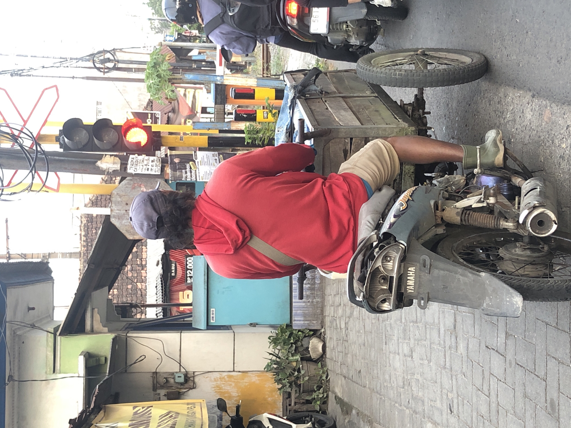In Sumorame, Indonesia, a man wearing a red hoodie and tan shorts rides a modified motorcycle that has been fitted with a wooden cart in the rear. He is wearing green rubber boots. The motorcycle is dark green and has "YAMAHA" written on the rear fender. The cart is made of weathered wood planks. A traffic light with a red signal is visible in the background. Other motorcycles and traffic signals are also present in the urban environment.