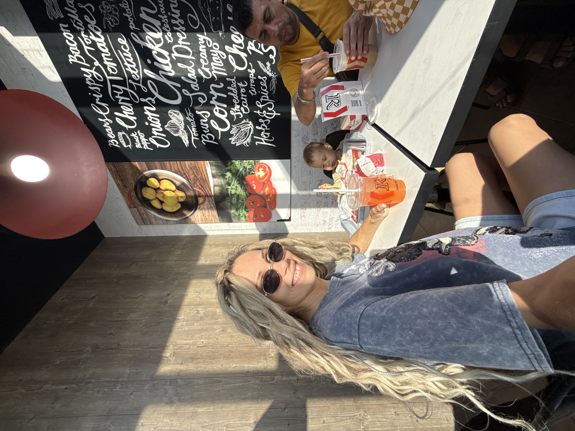 A woman with long, blonde dreadlocks and sunglasses smiles at the camera, seated at a white table. She wears a distressed grey t-shirt with a sequined graphic. To her right, a man in a yellow t-shirt holds a drink with a straw. A young child is seated in a high chair between them. On the table are several fast-food cups, bags, and what appear to be french fries. A chalkboard menu with white lettering listing food items is visible on the wall behind them. The scene is illuminated by natural sunlight casting shadows. The location is identified as Rusanovo, Russia.