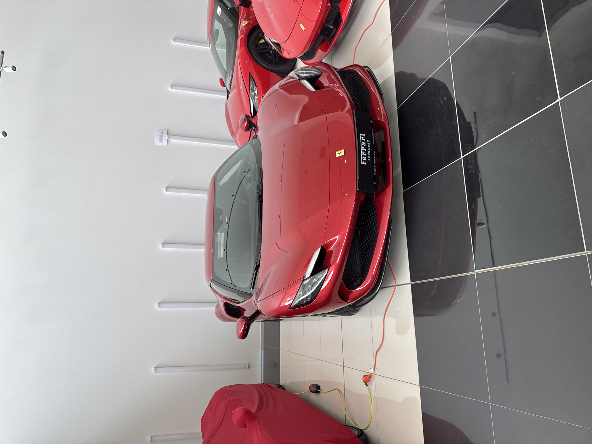 The image captures an indoor scene featuring multiple red Ferrari sports cars, likely within a showroom or private garage. The dominant car in the foreground is a red Ferrari, positioned diagonally and facing slightly towards the viewer. Its sleek, modern design is accentuated by its glossy finish, reflecting the bright overhead lighting.
On the front bumper of this central Ferrari, a black plate is visible, displaying white text: "Ferrari APPROVED" at the top, "Ayto Импълс" (which translates to "Auto Impulse") below it, and "www.ferrari.bg" at the bottom. A small, iconic Ferrari prancing horse emblem is also present on the car's hood.
Behind and to the left of the prominent Ferrari, another red Ferrari is partially visible, showing its front-end details. In the lower left corner of the frame, a third red car is seen covered with a red car cover, suggesting it's either stored or awaiting maintenance. A yellow charging cable with an orange connector runs from a power outlet on the wall towards the covered car, and another red cable extends from the foreground Ferrari, indicating that at least two vehicles are connected for charging or power.
The setting features clean, white walls illuminated by multiple horizontal linear lighting fixtures. The floor is composed of large, highly polished black and white tiles, creating strong reflections of the cars and the overhead lights. The overall ambiance suggests a high-end display or maintenance area for luxury vehicles. The image is captured with a vertical orientation (rotated 90 degrees clockwise from standard landscape). The location context is Sofia, Bulgaria, suggested by the "www.ferrari.bg" text.