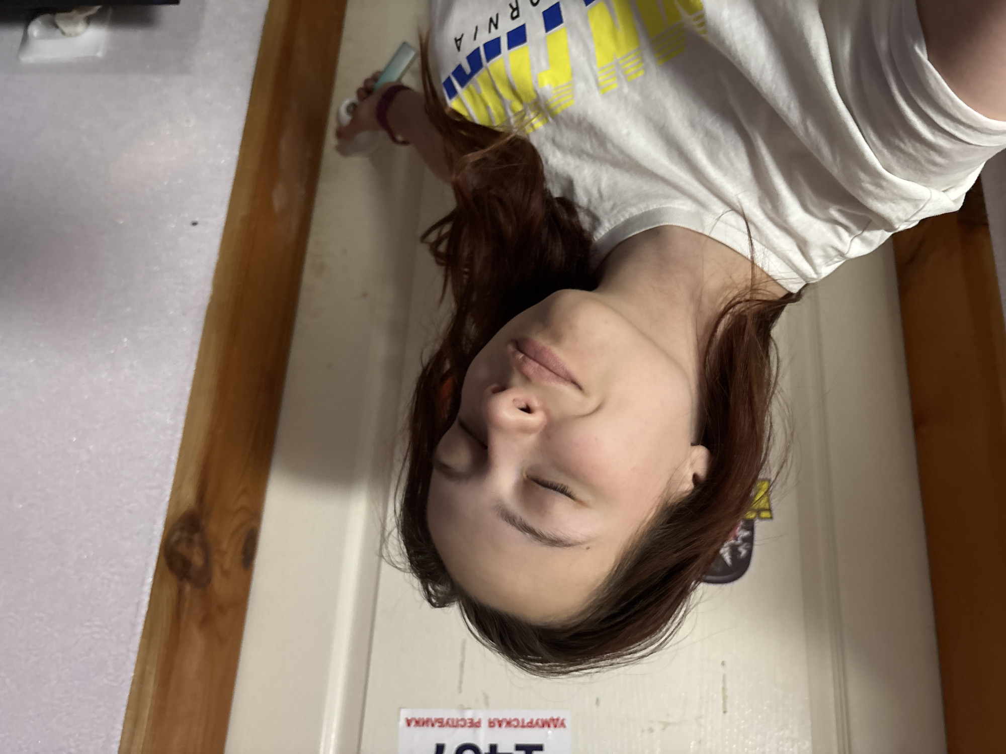 A woman with long brown hair is lying down, facing the camera. She is wearing a white T-shirt with yellow and blue stripes and the word "California" printed on it. She has her eyes closed. The background appears to be a white door with a wooden frame on the left side. On the door, there is a sign with text in Russian, including the words "ЯМАУРТСКАЯ РЕСПУБЛИКА". It is likely the image was taken in Прикамский, Russia.