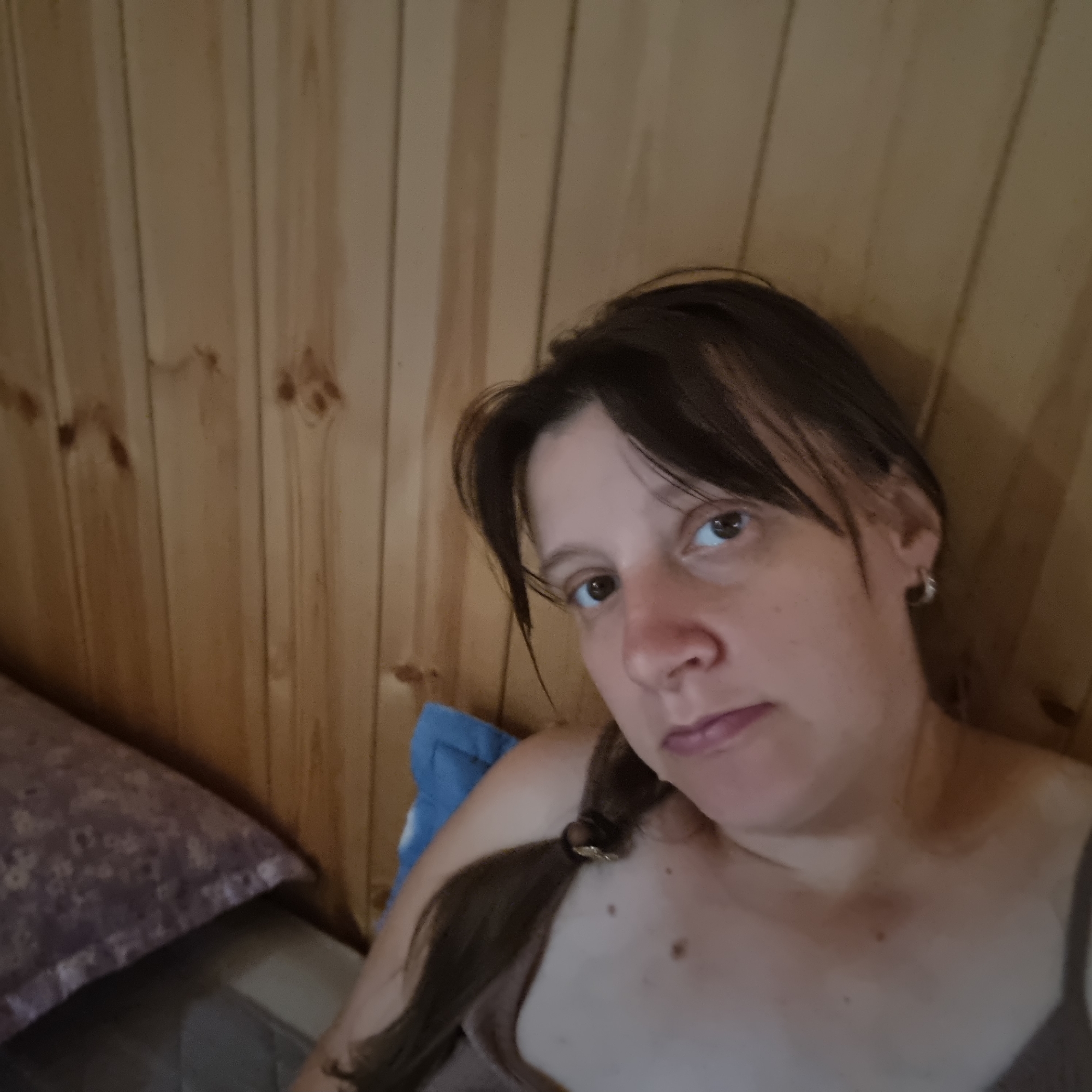 A medium shot of a woman with brown hair pulled back in a braid, lying down in front of a light brown wooden wall. She is wearing a tan colored top. A pillow with a floral pattern is to her left. The image appears to be taken indoors, possibly in a home setting. The location is not identifiable within Dunnae-myeon, South Korea from the image alone.