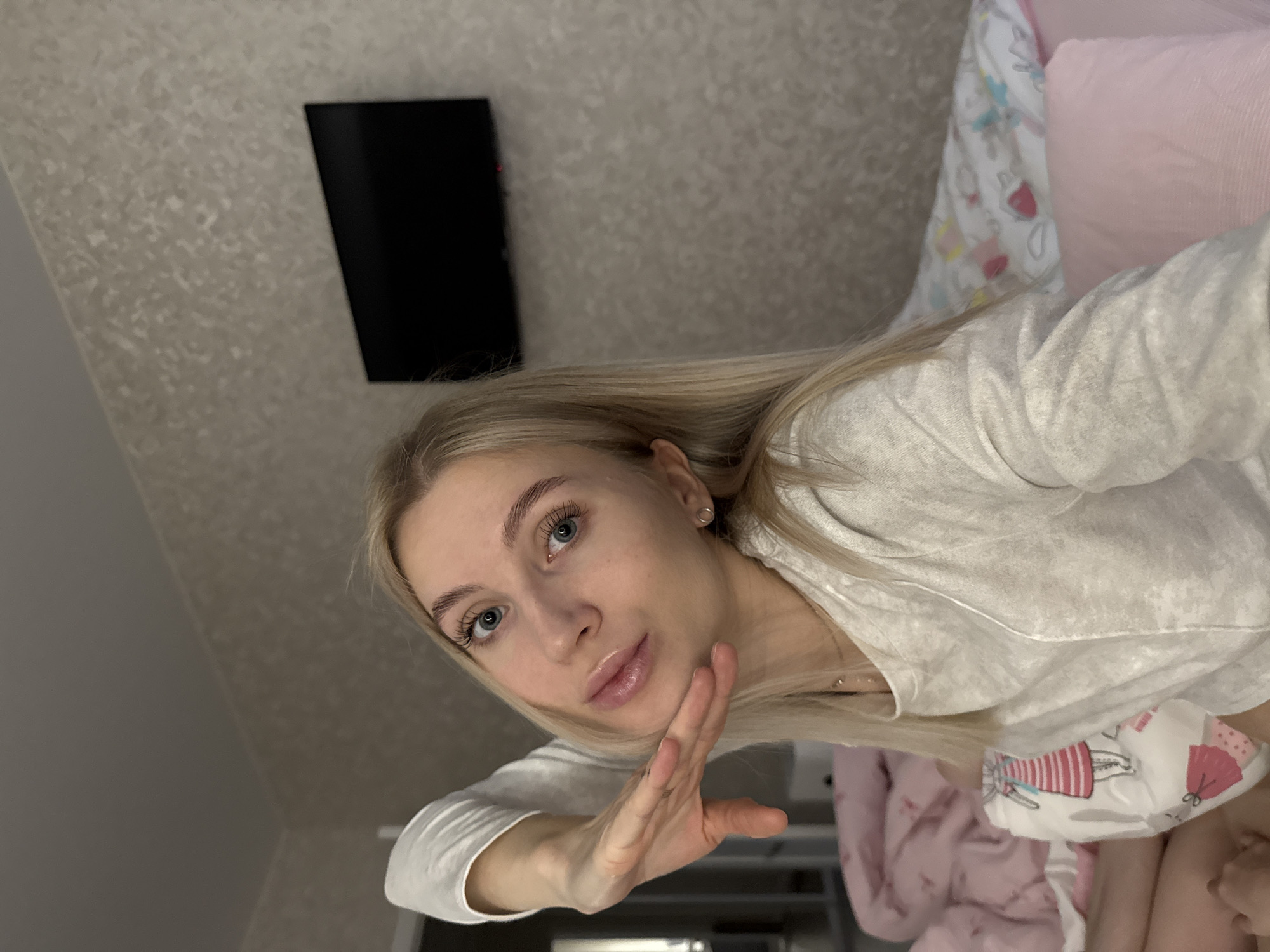 A young woman with light blonde hair and blue eyes is lying on a bed, likely indoors, with a wall-mounted television visible in the background. The bedding includes a pink pillow and a patterned sheet. The location is suggested to be Izhevsk, Russia.