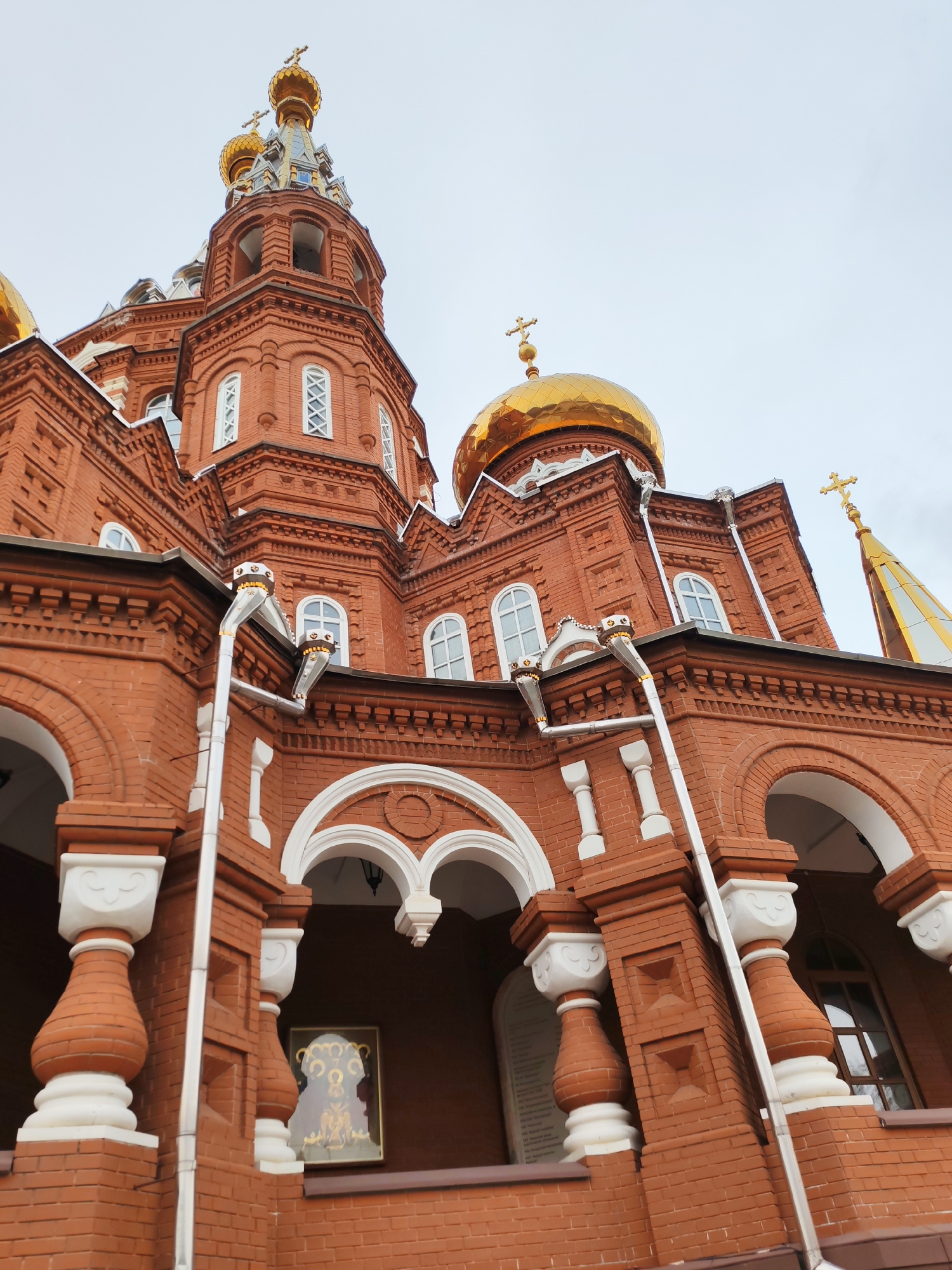 This low-angle shot captures a grand Russian Orthodox church, primarily constructed from warm red brick with ornate white architectural details and multiple shimmering golden domes. The building stands impressively against an overcast, light gray sky.
In the foreground, the church's lower facade features large arched entrances and windows. Sturdy brick pillars are adorned with decorative white balusters at their bases and tops. Under one of the main arches, a framed religious icon is displayed, depicting figures with golden halos and detailing. Another archway to the right appears to have a plaque with unreadable text.
The brickwork throughout the structure is intricate, featuring various patterns, cornices, and decorative elements. Numerous white-framed windows, many with arched tops and some with X-shaped cross-bars, punctuate the red brick walls. Several metallic rainwater downspouts, accented with small golden decorative elements, run vertically along the building's facade.
The upper sections of the church are crowned with multiple golden onion domes, each topped with a distinct golden Orthodox cross. A tall, central bell tower or cupola rises towards the sky, featuring arched openings and a golden spire capped with a cross. To its left, a larger golden dome is partially visible, contributing to the church's majestic silhouette. The overall design reflects traditional Russian ecclesiastical architecture. The cloudy sky suggests a soft, diffused light, enhancing the vibrant colors of the brick and the reflective quality of the gold. This detailed view showcases the intricate craftsmanship of the church, located in Izhevsk, Russia.