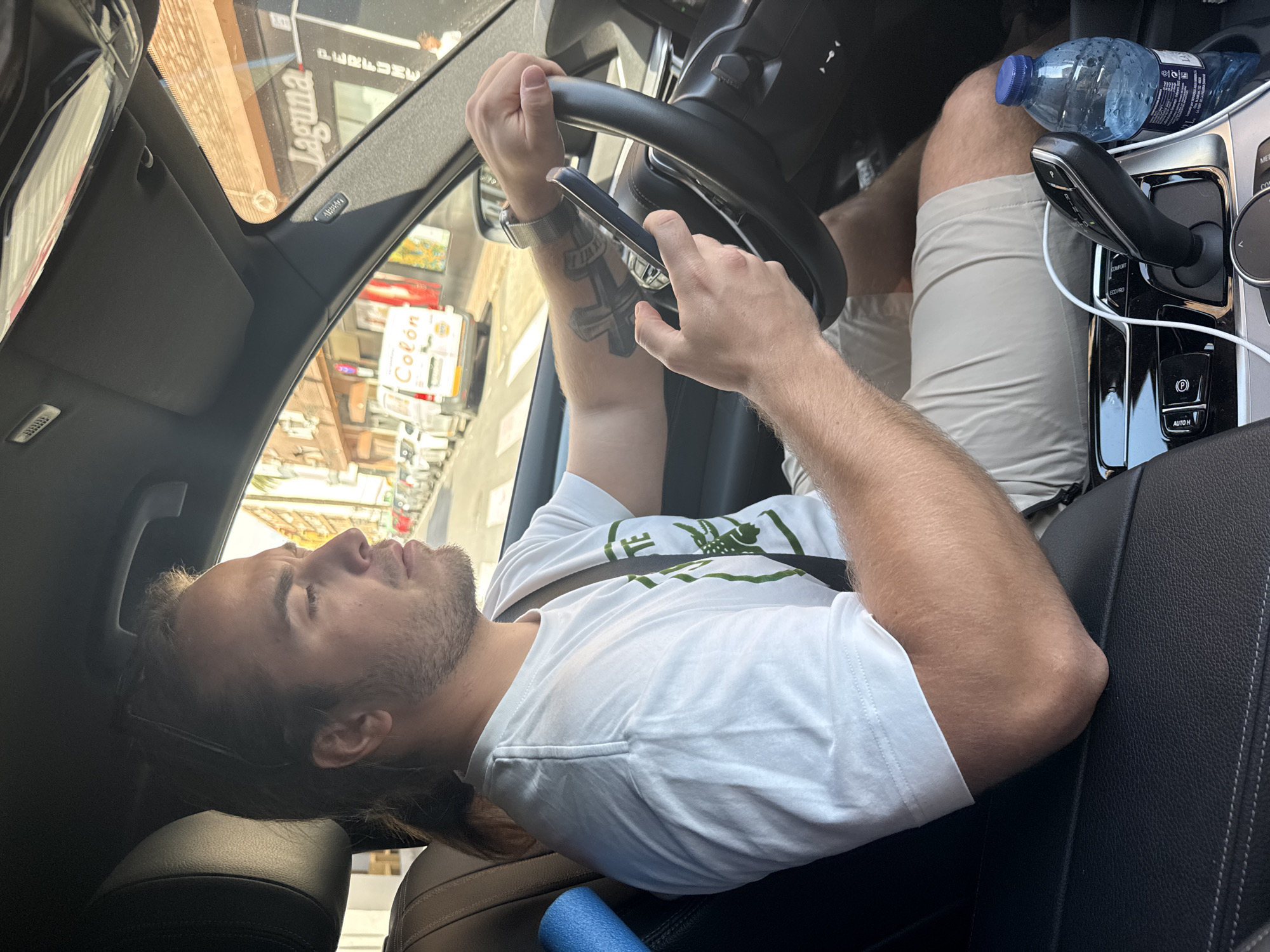 The image shows the interior of a car on a bright, sunny day, with a focus on the driver and the street outside. A light-skinned man with dark hair (possibly tied back) and stubble is seated in the driver's seat. He wears a white short-sleeved t-shirt with a green graphic and light-colored shorts. A prominent cross-like tattoo is visible on his left forearm, and he wears a smartwatch on the same wrist. Sunglasses are pushed back on his head.
The man is in a relaxed, almost lounging position, with his right leg bent and propped up. He holds a smartphone in his right hand, while his left hand rests on the steering wheel. The car's interior features a black steering wheel, an automatic gear shifter, and a center console with buttons labeled "COMFORT," "ECOPRO," and "AUTO H," as well as a "P" for the parking brake. A blue water bottle and a white charging cable are in the console. Dark leather seats are visible, with a blue foam roller resting on the passenger side.
Through the windshield, street buildings and reflections are visible. The word "Laguna" appears reflected upside down, and a sign with "Colón" can be seen outside. "AIRBAG" is visible on the car's A-pillar. The scene depicts a calm moment, likely while the car is stationary. Based on the "Colón" sign and provided context, the location is Valencia, Spain.