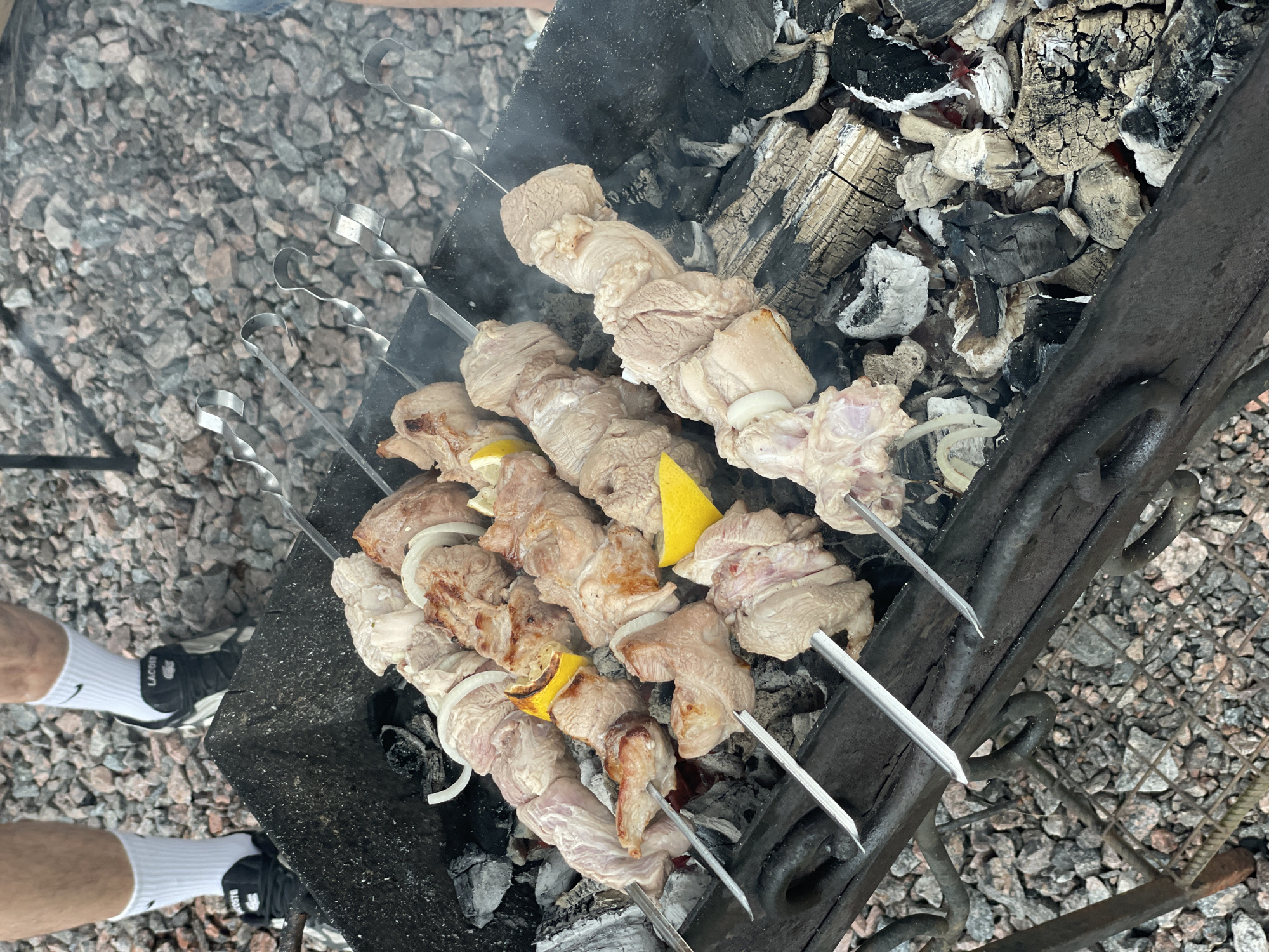 The image shows a close-up view of several skewers of raw meat interspersed with onion slices and lemon wedges, being cooked over hot coals in a black metal brazier. The brazier is placed on a bed of grey gravel. In the background, partially visible legs of individuals wearing white socks and dark sneakers are seen, standing on the gravel. The scene suggests an outdoor barbecue or gathering. The location is identified as Novo-Tuutari, Russia.