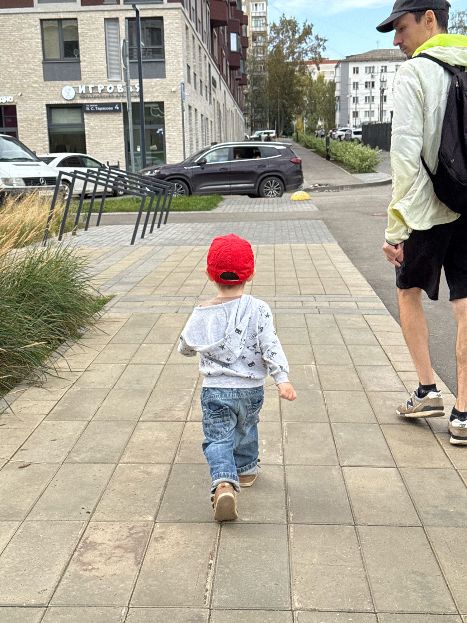 The image shows a child wearing a red cap and blue jeans walking away from the camera on a paved sidewalk. The child is wearing a gray hoodie with a star pattern and brown shoes. To the right of the child, an adult male wearing a black baseball cap, a light-colored jacket with a neon green collar, black shorts, and athletic shoes is also walking. The adult is carrying a black backpack. In the background, there is a modern brick building with windows and signs in Russian, one of which reads "ИГРОВЫЕ" (Igrovye), indicating a gaming or entertainment establishment. Cars are parked on the street, and there is a bicycle rack in the foreground. The scene is set in a city environment with a cloudy sky. The building in the background has the address "ул. С. Тарасова, 4" (S. Tarasova St, 4) visible, which confirms the location as Izhevsk, Russia.