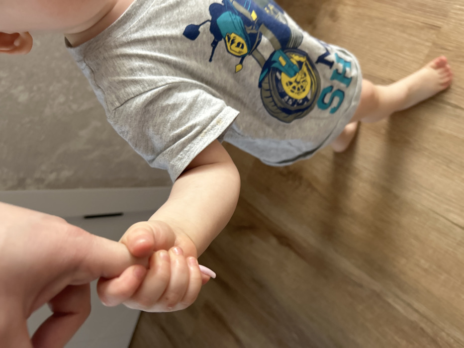 An adult's right index finger, with light pink nail polish, is gripped by the left hand of an infant in an indoor setting. The infant's left arm and part of its torso are visible, wearing a grey short-sleeved t-shirt featuring a blue, yellow, and black motorcycle graphic and the word "SHARK" in blue capital letters. The infant's bare feet are positioned on light brown wooden flooring, suggesting a standing or walking posture. A light-colored, textured wall and a segment of a white doorframe are visible in the background. The specific location within the city of Chaikovsky, Russia, cannot be determined from the visual evidence presented in the image.