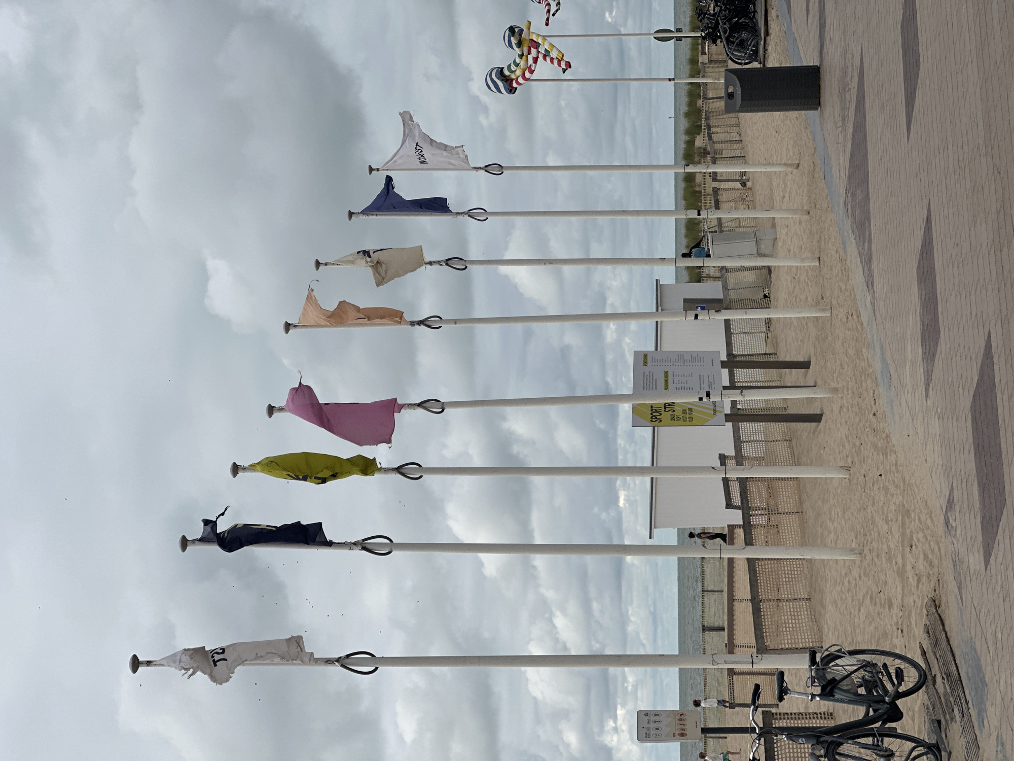 The image depicts a cloudy day at the beach in Knokke-Heist, Belgium. In the foreground, a brick walkway lines a sandy beach. To the left of the walkway, a black bicycle is parked on a wooden plank. A row of white poles with flags extends along the beach. The flags vary in color and design; some are plain, while others have text or patterns. Behind the poles, a low fence separates the walkway from the sand, and beyond that, a building with a white exterior is visible. The sea is in the background, with a hazy horizon under a grey, overcast sky. Several people are present on the beach, including a few individuals walking near the fence and by the water's edge. To the right, a dark trash can and a cluster of parked bicycles are visible near the end of the walkway. Further down the beach, a whimsical, striped flag resembling an octopus or jellyfish on a pole is also present.