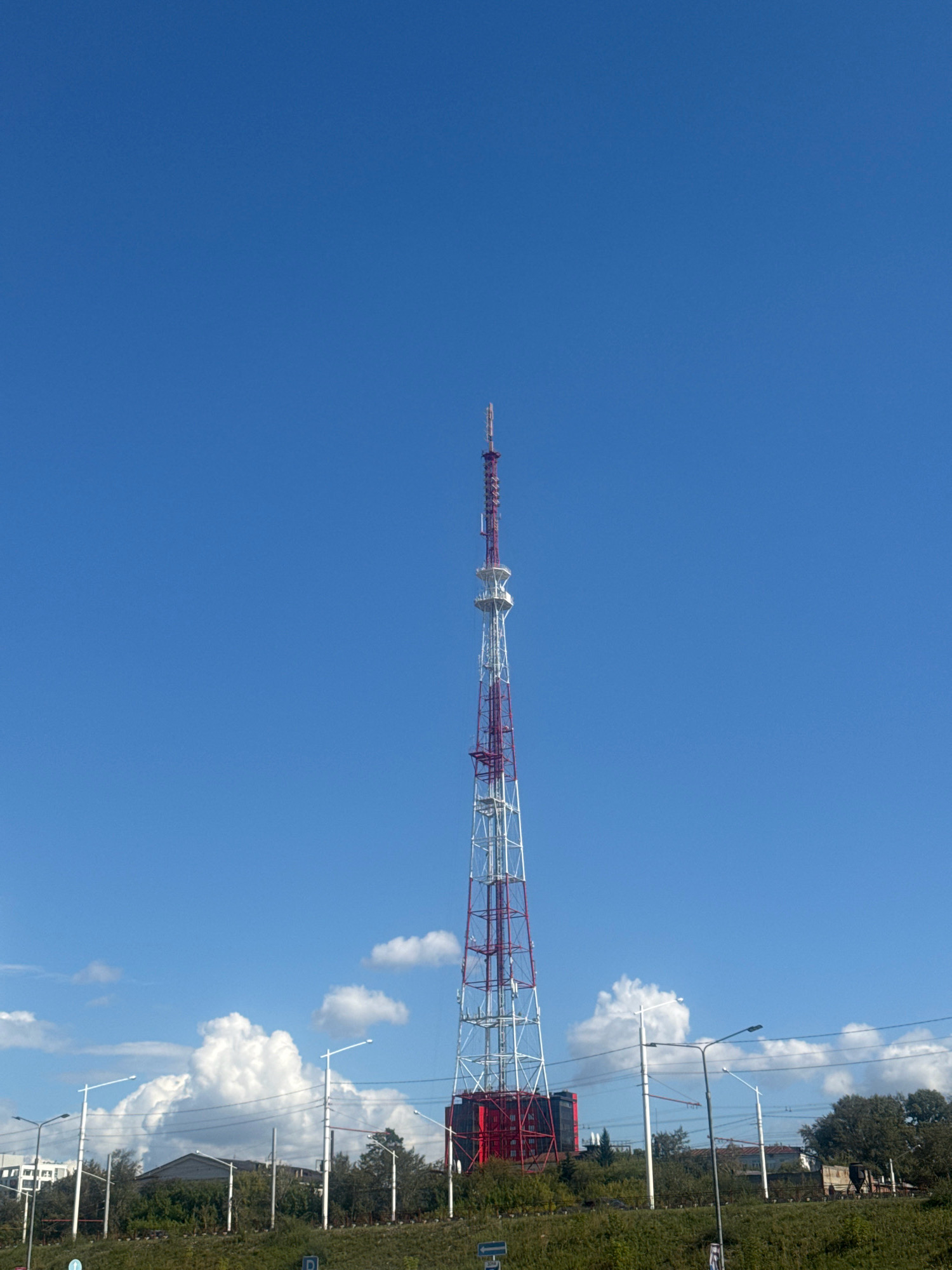 An outdoor, low-angle, eye-level, medium shot captures the Irkutsk Television Tower, a tall, red and white lattice structure, dominating the center of the frame against a clear, bright blue sky. The tower tapers towards the top, with various antennas and equipment visible. At the base of the tower, a red building complex is situated. In the foreground, a grassy slope is covered with green foliage. Behind the slope, a line of streetlights and utility poles with power lines runs horizontally across the middle ground. In the distance, several buildings and trees are visible, suggesting an urban or semi-urban environment. Puffy white clouds are scattered across the sky, mostly concentrated on the left side of the frame. The lighting suggests it is daytime, likely late morning or early afternoon, with the sun casting a slight shadow. There are no people visible in the image.