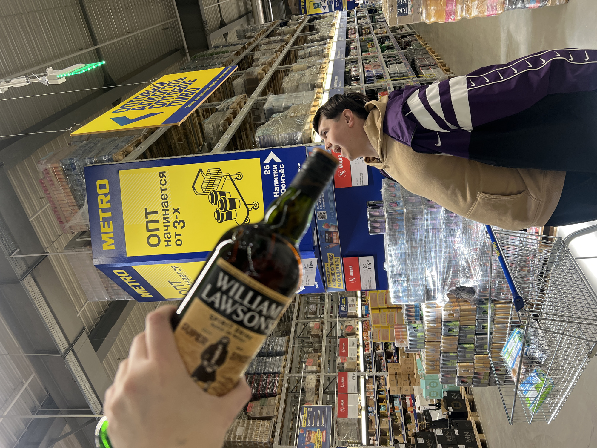 The image captures a moment inside a wholesale grocery store, most likely a METRO Cash & Carry, judging by the signage. In the foreground, a hand holds a bottle of William Lawson's Scotch whisky. The subject is centered in the frame. In the middle ground, a young man, wearing a beige hoodie under a purple Nike jacket, looks towards shelves of products. A shopping cart is partially visible. The background features high shelves stacked with assorted goods, typical of a wholesale environment. Russian language signage is visible, confirming the location in a Russian speaking country. It is difficult to pinpoint that exact location in Izhevsk without additional identifiers.