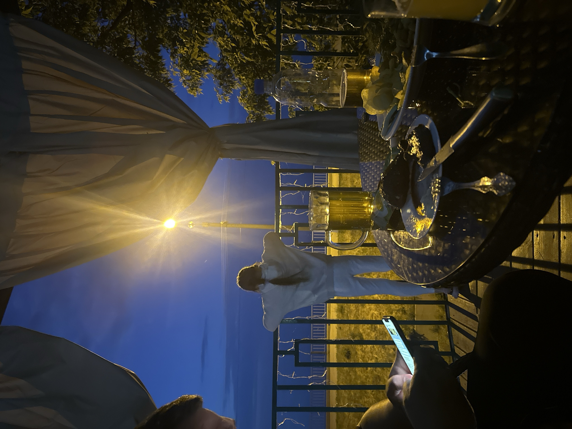 An outdoor dining area at dusk, possibly Khvalynsk, Russia, is depicted. A young person wearing a white tracksuit stands by a railing, facing away from the camera and looking out at the darkening sky illuminated by a bright lamp. On a glass-top table in the foreground, there are plates of food, cutlery, and several glasses of beer. One person is visible in the lower left corner, engaged with a smartphone. White curtains hang from an overhead structure, and trees are visible to the right. The scene is lit by artificial light from a lamp and the ambient twilight.