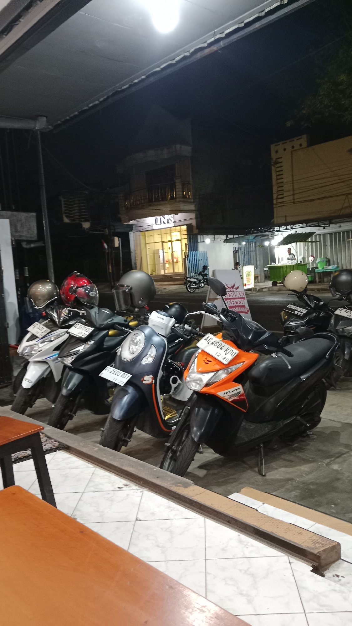 The image depicts a nighttime street scene in Blitar, Indonesia, with a row of parked motorcycles in the foreground. Several helmets are placed on the seats of the motorcycles. In the background, a building with illuminated signage that reads "UNS" is visible, suggesting a commercial establishment. The street is paved with tiles, and a portion of a wooden table is visible in the lower left corner. The overall environment appears to be an outdoor area adjacent to a building, likely a cafe or shop, with a few people visible further in the background, one of whom appears to be working at a food stall. The lighting suggests it is nighttime, with artificial lights illuminating the scene.