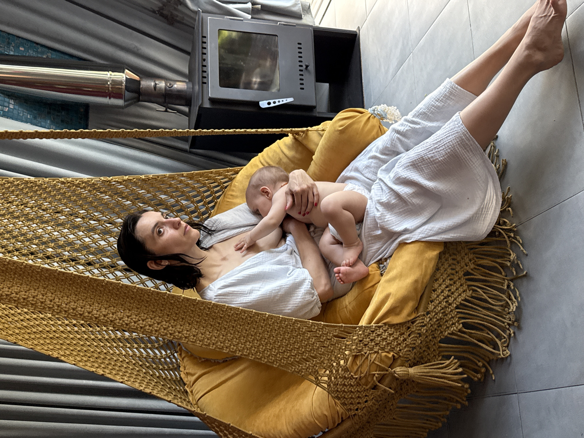 A woman lies in a yellow macrame hammock indoors, breastfeeding a baby. She wears a white garment, and her dark hair is damp. The baby is partially unclothed. A black stove sits to the side, with a metal pipe extending towards the left. The hammock is suspended indoors, and the floor is tiled. The location provided, Чёрное, Russia, cannot be verified.