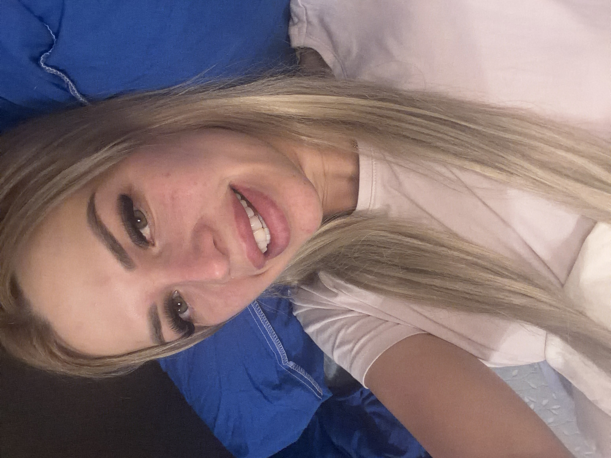 The image shows a close-up, overhead shot of a young Caucasian woman with blonde hair, lying on a bed with blue pillows. She is wearing a light pink t-shirt. Her eyes are open and looking towards the camera, and she is smiling, revealing white teeth. Her makeup includes noticeable false eyelashes and lip gloss. The background is blurred, with the blue of the pillows and a hint of dark fabric. There are no discernible architectural features or environmental cues that would suggest a specific location within Kyiv, Ukraine. The scene appears to be an informal, personal setting.