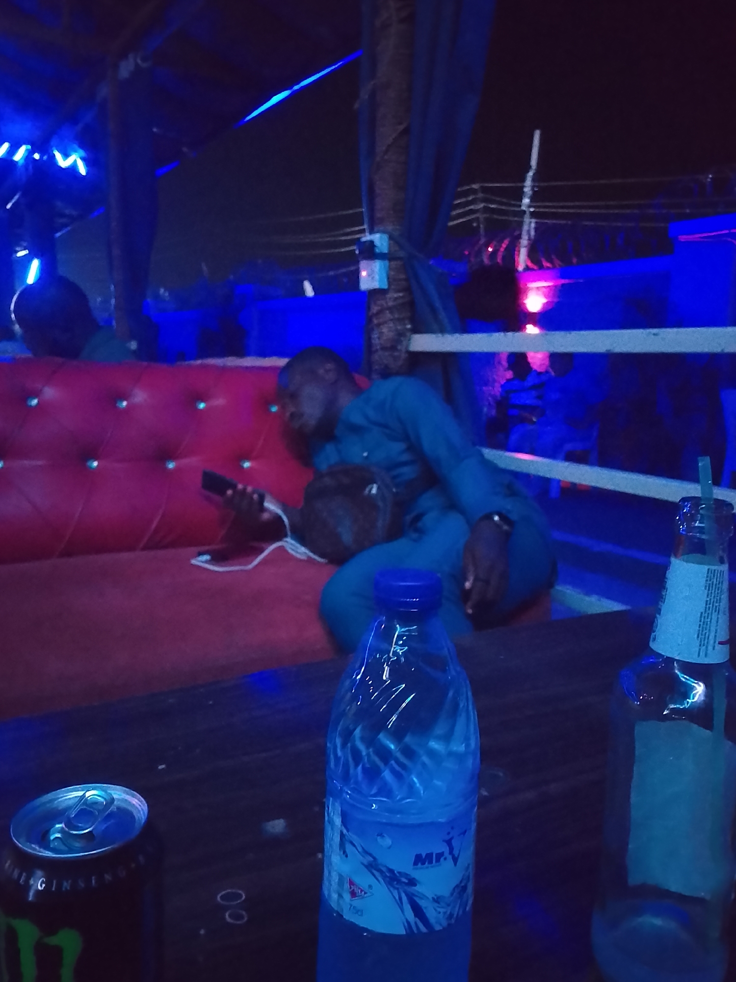 The image depicts a dimly lit outdoor area, likely a bar or lounge, in Narayi, Nigeria. A man in blue clothing reclines on a red tufted sofa, holding a mobile phone and a brown object, possibly a bag or wallet. On the table in front are a can of Monster energy drink, a bottle of water labeled "Mr. V", and an empty bottle with a straw. The scene is illuminated by blue and purple lights, and elements of the outdoor structure are visible in the background.