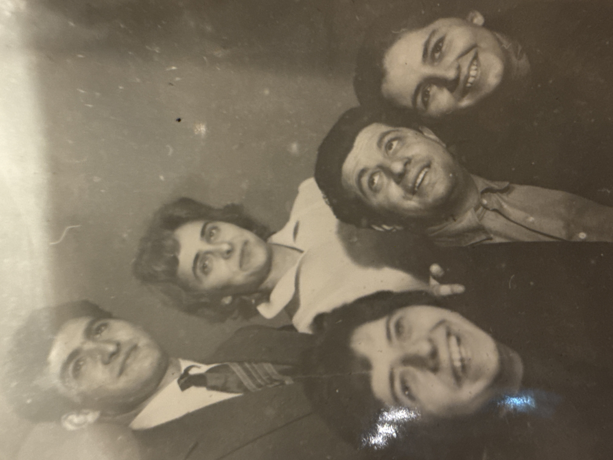 The image is a faded black and white photograph depicting five people. From the bottom left, a man in a suit and tie looks upward. Next to him, a woman with curly hair looks towards the viewer. Above her, a man in a button-down shirt also looks upward. At the top right, a woman smiles at the viewer. Below her, another woman with dark hair smiles broadly. The background is indistinct and appears to have marks and fading common in old photographs. The location is not discernible from the image. No specific actions or interactions are evident other than the subjects posing for the photo.