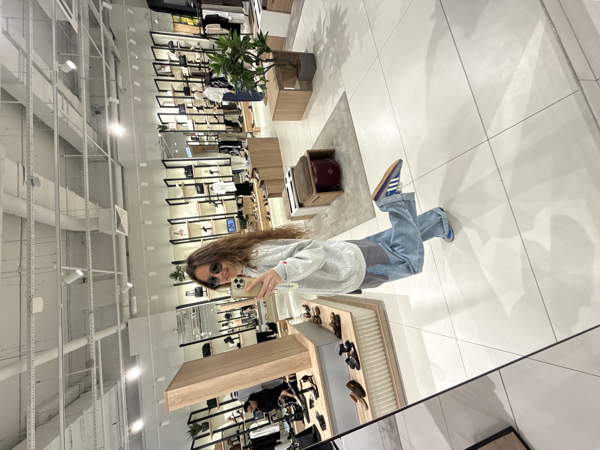 A woman stands in a brightly lit, modern retail store, possibly a boutique or department store, in Moscow, Russia, taking a selfie in a large mirror. She has long, wavy brown hair and wears sunglasses, a light grey crewneck sweatshirt over a darker grey undershirt, and loose-fitting light blue jeans. Her left leg is bent and raised, showing off her colorful striped athletic shoes. The store features shelves stocked with handbags, shoes, and clothing. In the background, other shoppers can be seen browsing. The store's interior is characterized by white tiled floors, white ceilings with visible ventilation and lighting fixtures, and wooden display units. A potted plant is visible on a wooden stand to the right. The overall impression is one of a clean, organized, and contemporary retail environment.