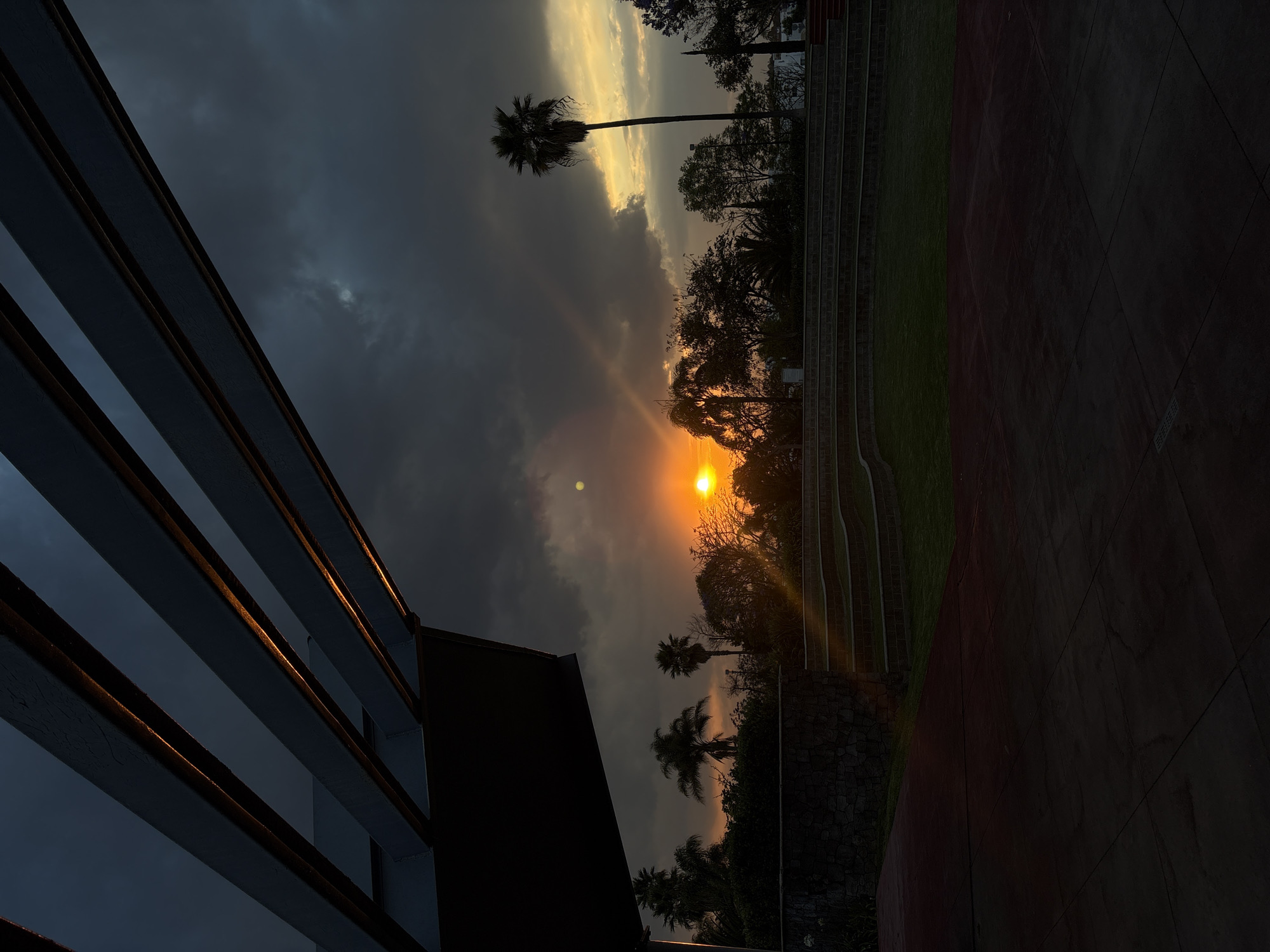 The image captures a dramatic sunset scene over an outdoor area in San Bernardino Tlaxcalancingo, Mexico. The sky dominates the upper portion, featuring a striking contrast between dark, heavy clouds and bursts of golden light. The sun, a bright orange orb, is visible on the right side, casting a warm glow and creating a sunbeam effect towards the ground.
To the left, a dark modern building or structure with prominent horizontal slats is partially visible, its edges catching the golden reflections of the setting sun. These architectural elements are silhouetted against the moody sky.
The middle and right sections of the image are filled with a variety of trees, including several tall palm trees, which appear as dark silhouettes against the vibrant sky. Below the trees, the landscape includes green grass and terraced stone elements on the right, leading down to a reddish-brown paved area or path. The lighting suggests late afternoon or early evening, creating a serene yet powerful atmosphere. There is no discernible human activity or visible text in the image.