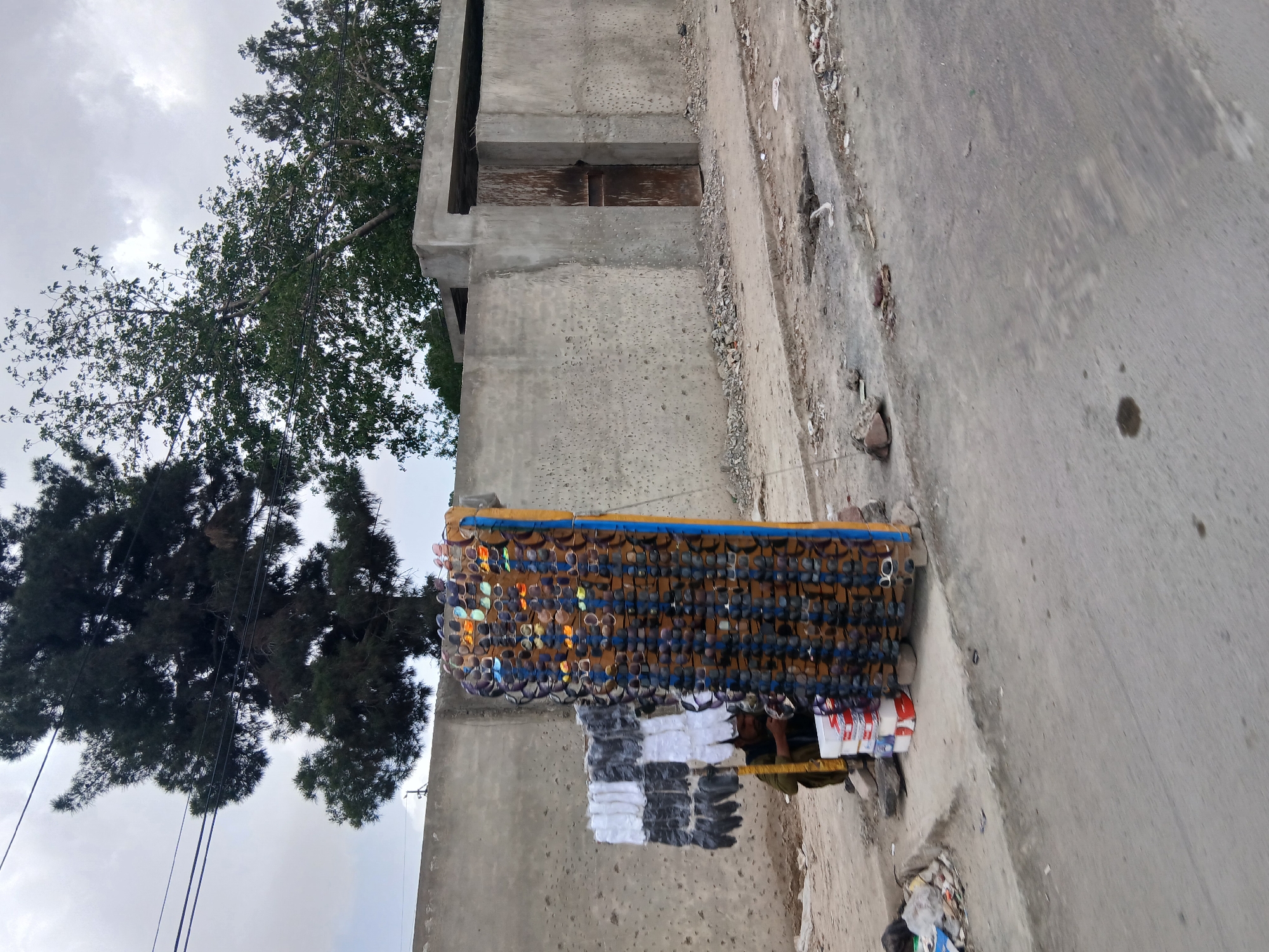 The image shows a street scene in Quetta City Tehsil, Pakistan. A vendor displays a large collection of sunglasses on a wooden rack beside a concrete building. Below the sunglasses, a display of white and gray items (possibly clothing) is visible. A man is present behind the display. The scene is next to a street. Trees are visible.