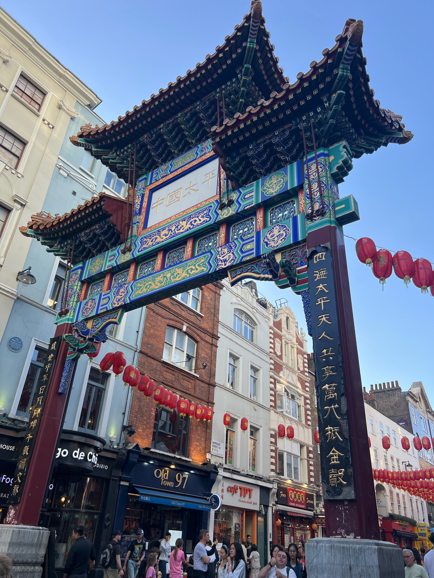 The image shows the archway in London's Chinatown, situated in the West End of the city. The archway is ornately decorated in traditional Chinese style with a dark red base, intricate blue and gold detailing, and a layered, tiled roof. Chinese characters are visible on the archway and a tall column beside it. Numerous red lanterns are strung across the street, hanging from wires and poles. The background consists of buildings with a mix of architectural styles, some appearing older and others more modern, with shopfronts at street level. The street is populated with a number of people walking and gathered. The sky above is clear and blue, indicating daytime.