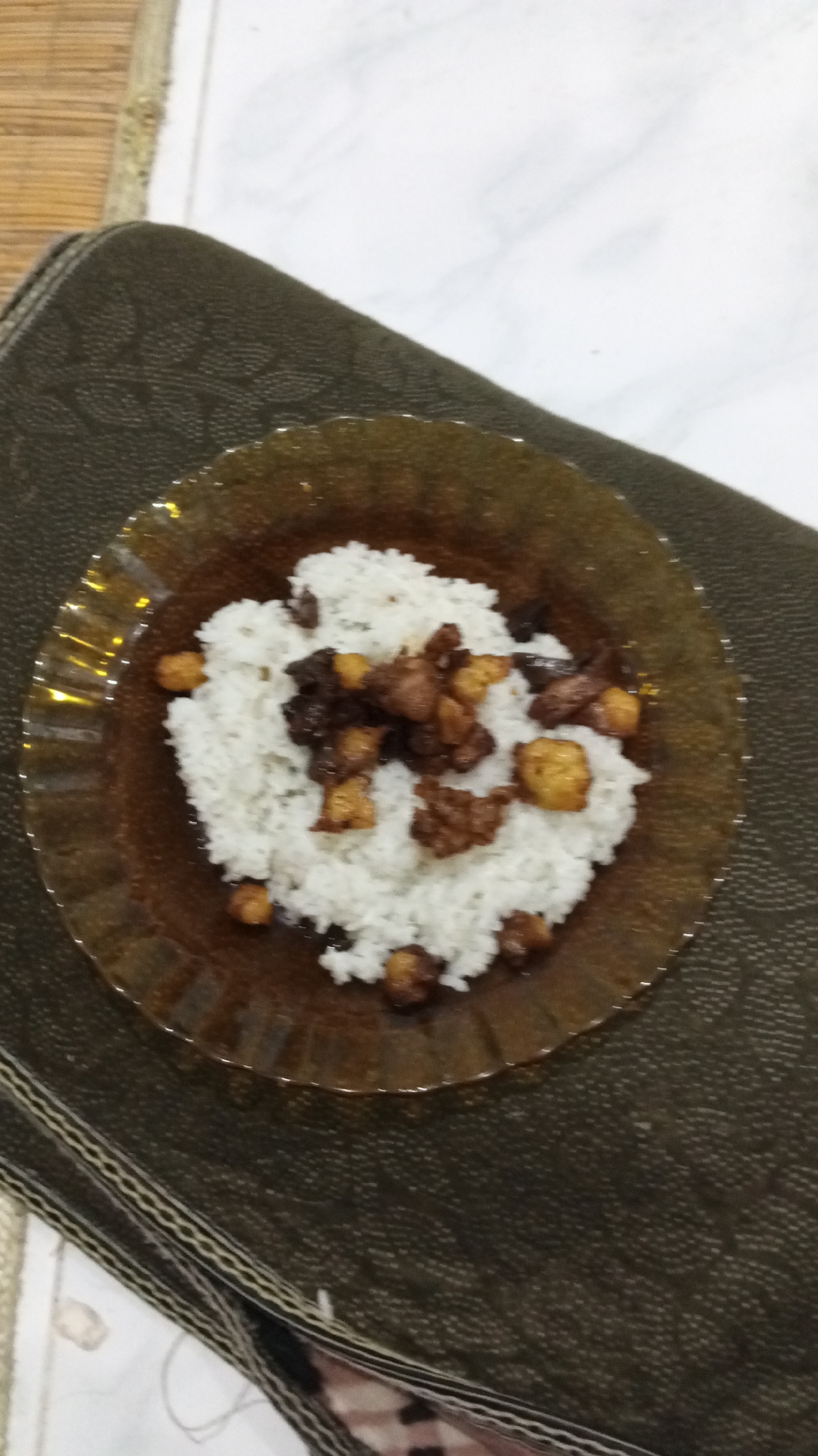 A plate of white rice with fried tempeh and a dark sauce rests on a patterned cushion. The cushion is on top of a table. The table is indoors. The location is Lhokseumawe, Indonesia.