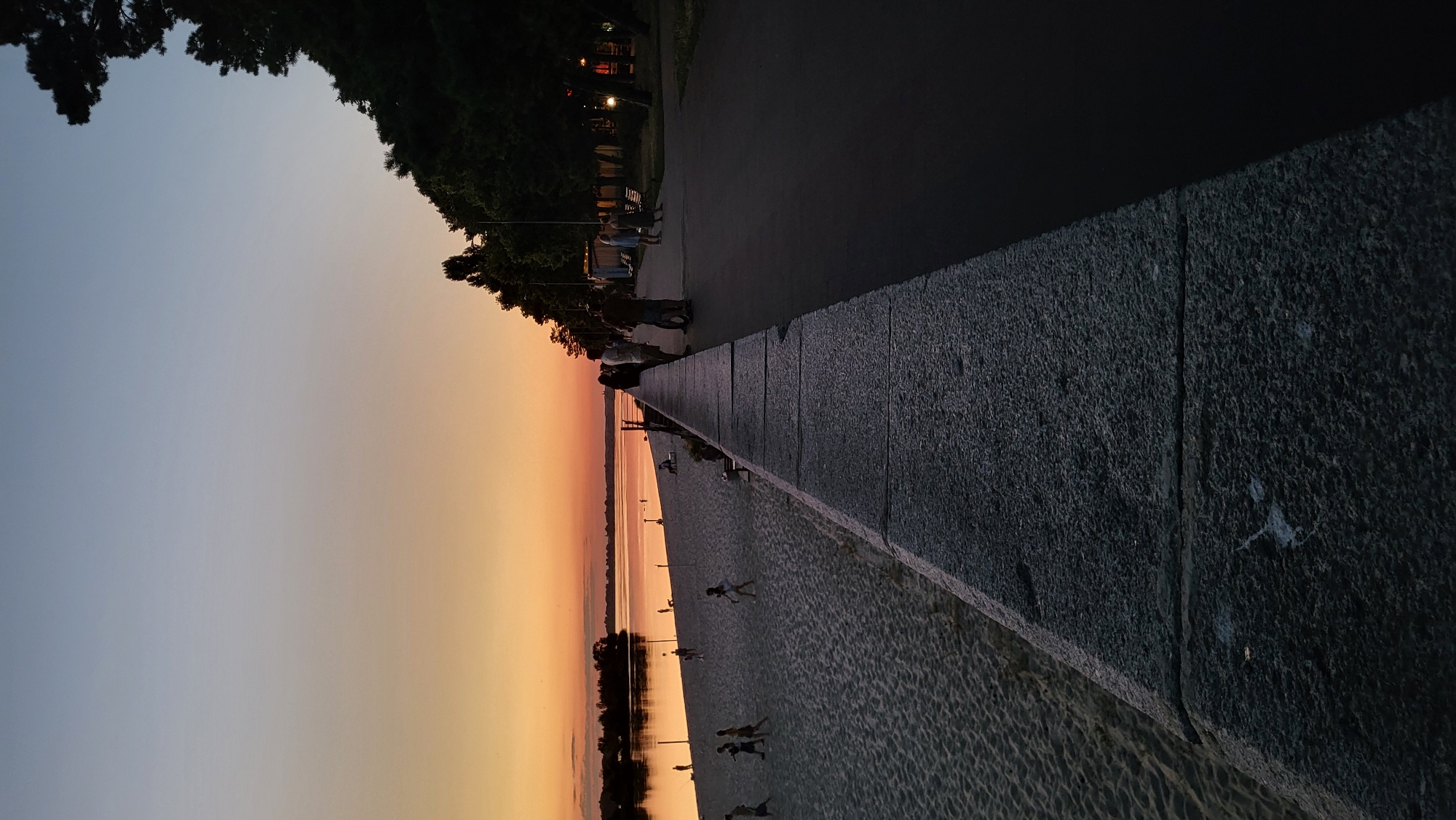 The image depicts a sunset scene along a waterfront. On the left side, a sandy beach is visible with several people playing or walking. Volleyball nets are set up on the sand. The water stretches out to the horizon, reflecting the warm hues of the sunset. A wide, stone embankment separates the beach from a paved walkway on the right. This walkway is lined with trees, and a few people are standing and conversing. The sky transitions from a soft orange near the horizon to a pale blue overhead. The lighting suggests it is dusk, with the right side of the image becoming darker due to the setting sun.