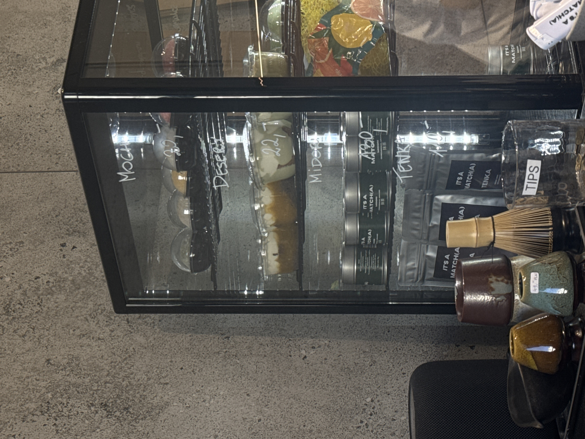 This image captures an indoor retail environment, likely a cafe or specialty shop, showcasing a variety of food and beverage-related products. The primary subject is a black-framed glass display case, presented at an angle where the grey, speckled concrete or terrazzo floor appears on the left side of the frame.
Inside the illuminated display case, various items are neatly arranged. On the far left, products are labeled "MOCHA" and "DESSERT," suggesting individual portions of sweet treats or drinks. Further to the right, a section includes an item with a label indicating "22," possibly a price. The middle of the case displays items associated with the label "MIDORI." The right side of the display is dedicated to matcha tea products, including cans and pouches. Visible text on these items includes "IT'S A MATCH(A)," "MIDORI," "430," "TENKA," and the Japanese characters for matcha (抹茶).
Outside and to the right of the display case, additional merchandise is arranged. A clear glass container prominently labeled "TIPS" is visible, likely for customer gratuities. Beside it, a traditional bamboo matcha whisk (chasen) is displayed along with several small, unique ceramic cups or bowls in earth tones like brown, teal, and mustard yellow. One teal cup features a price tag stating "49, PLN," which, along with the location context, identifies the currency as Polish Złoty. A part of a black mesh chair or similar object is also visible in the bottom right corner.
The scene depicts a static product display designed for retail, with no people present or explicit activity taking place. The indoor setting and visible pricing indicate a commercial establishment during operational hours.