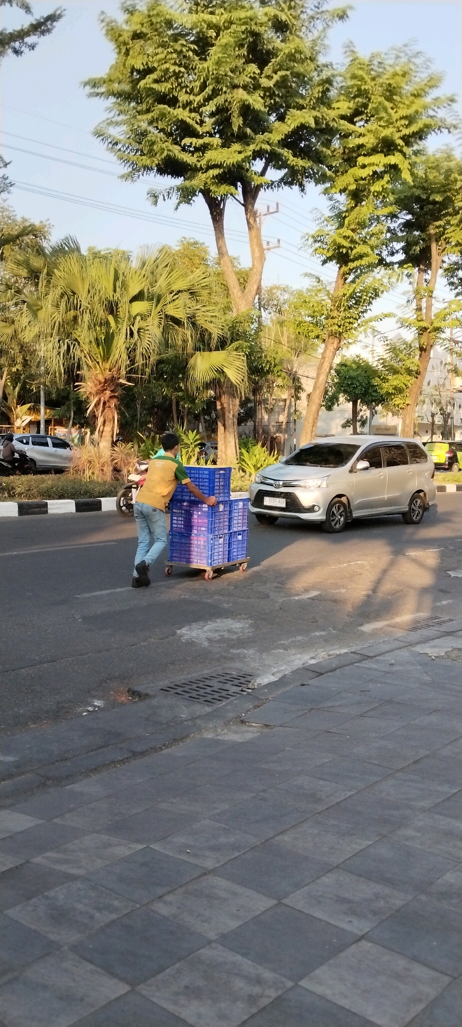 A lone man pushes a cart loaded with blue plastic crates along a street in Surabaya, Indonesia. He is wearing a yellow t-shirt and light blue jeans. The street is paved with asphalt and bordered by a sidewalk with a checkered pattern. Trees, including palm trees, line the street, and power lines are visible in the background. A silver car drives by in the opposite direction, and a motorcycle is parked on the side of the road. The sun is casting long shadows, suggesting it is either early morning or late afternoon.