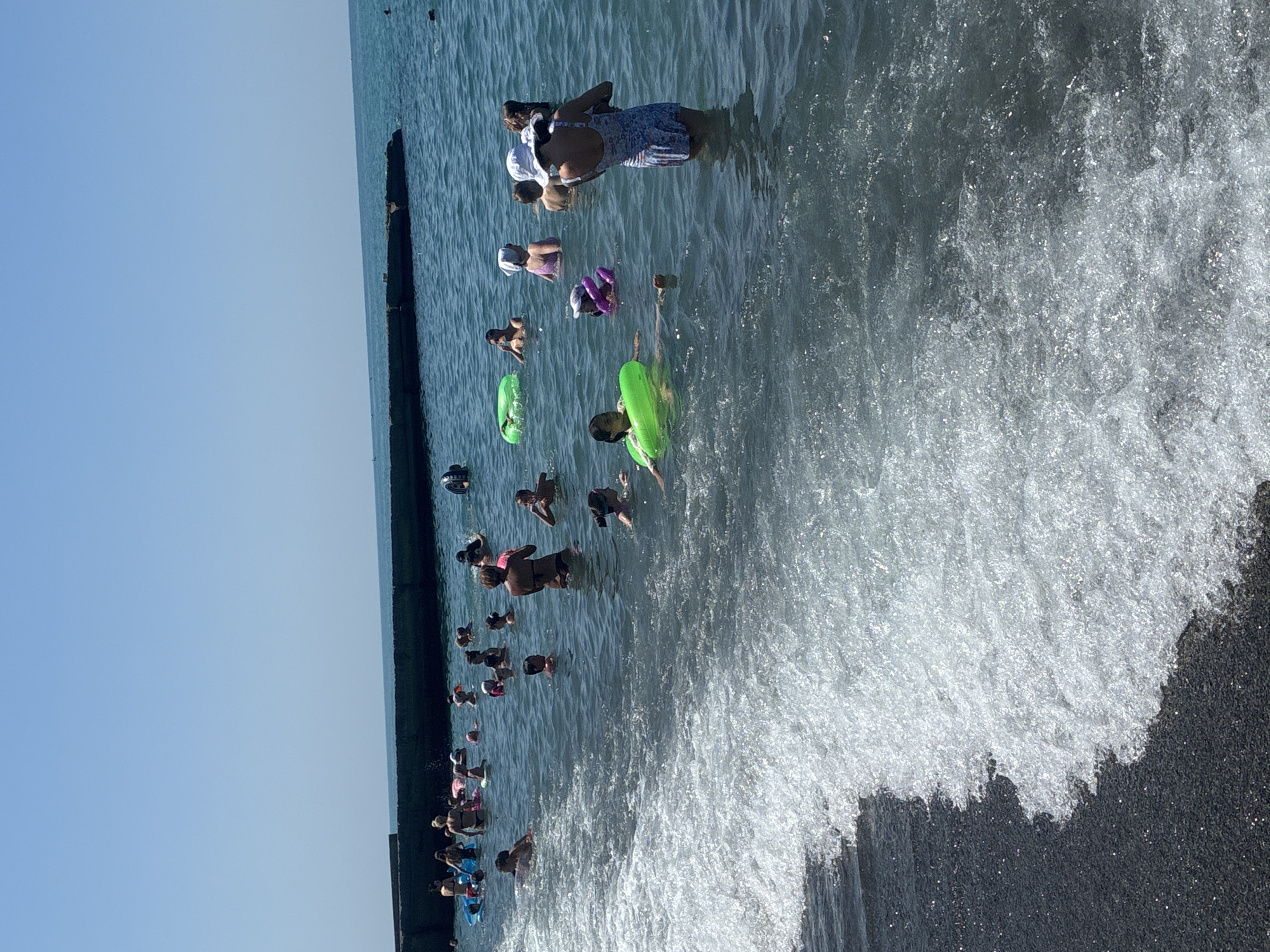 The image depicts a crowded beach scene at the city of Sochi, Russia, under a clear blue sky. The sea is a vibrant turquoise, with gentle waves lapping at the shore. A long, dark, concrete breakwater stretches across the background, with several people standing or sitting on it. In the water, numerous individuals, including adults and children, are engaged in various activities. Some are swimming, while others are relaxing on inflatable rings and rafts. In the foreground, the dark, pebbly shore is partially covered by a foamy wave. The overall atmosphere is one of lively recreation and enjoyment of a sunny day at the beach.