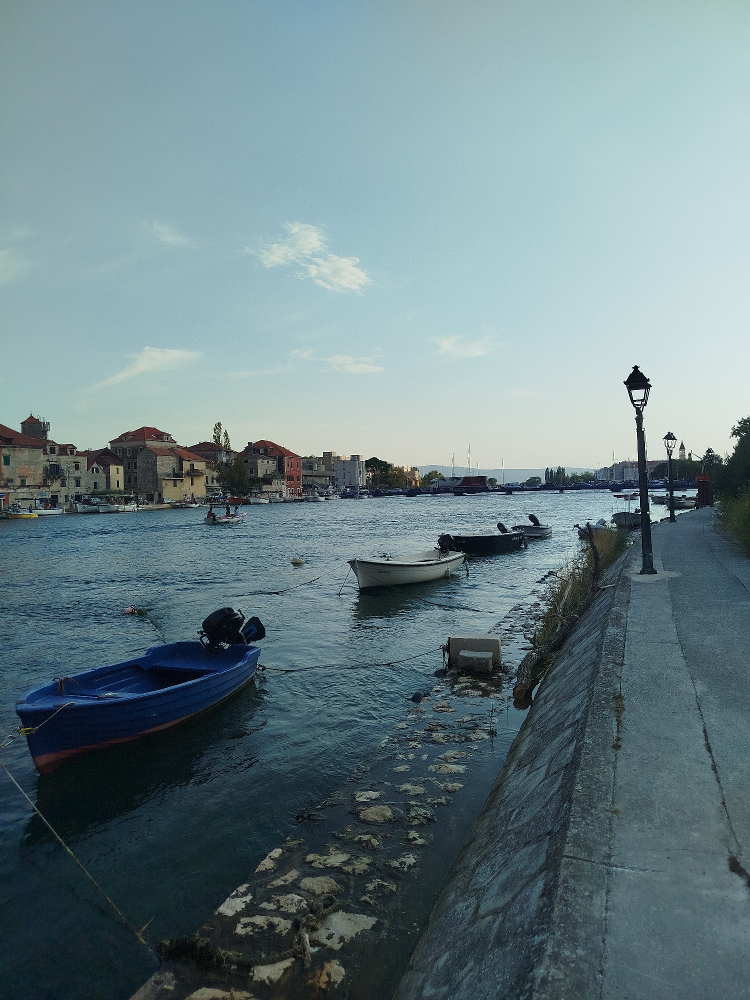 The image depicts a waterfront scene in Omiš, Croatia. A blue boat with an outboard motor is moored in the foreground. Several other boats are visible in the river, with a row of buildings lining the opposite bank. A paved walkway runs along the right side of the image, illuminated by a vintage-style lamppost. The sky is clear with scattered clouds.