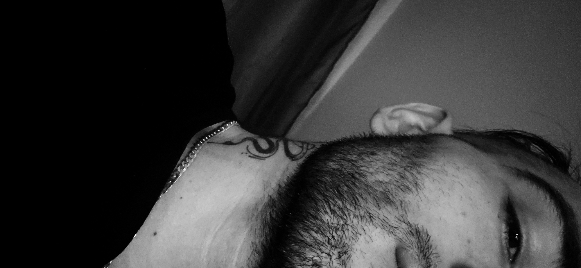 A close-up, monochrome image features the upper part of a male figure lying down. The man has a beard. His head is oriented sideways, with one eye visible. An ear is partially visible at the top of the frame. A shoulder with a tattoo is visible, along with a chain. A dark fabric is positioned adjacent to the shoulder. The background is gray. It is not possible to determine the location as Plovdiv, Bulgaria, as the environment is ambiguous.